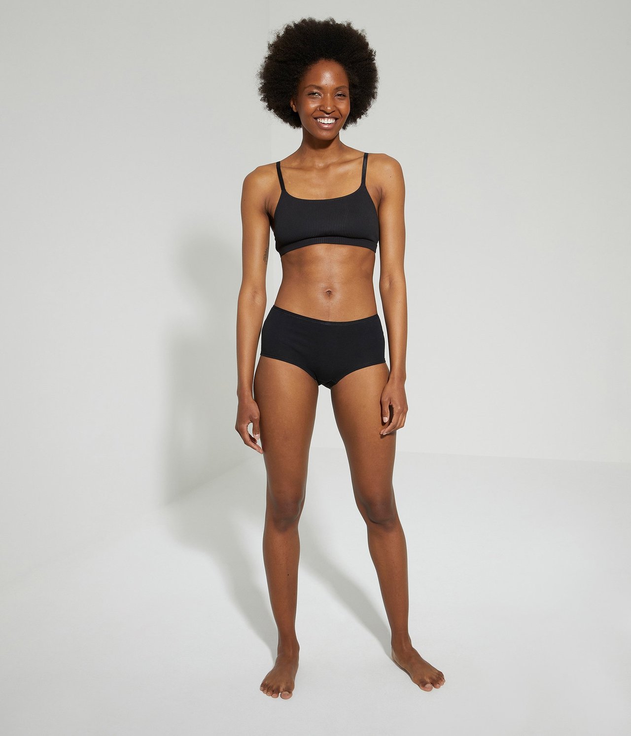 Widok z przodu: kobieta w czarnych bawełnianych majtkach typu hipster i czarnej prążkowanej braletce.