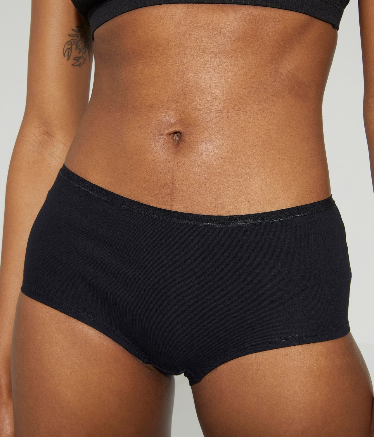 Zbliżenie z przodu kobiety w czarnych bawełnianych figach typu hipster i czarnym biustonoszu w prążki.