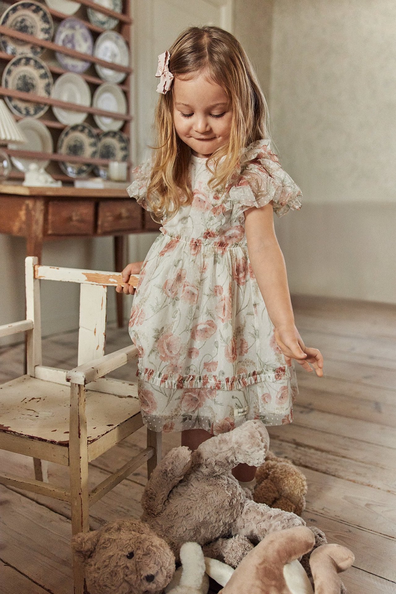 Framifrån: Barn i benvit blommig meshklänning med volangärmar, volangkant, och rosa hårrosett.