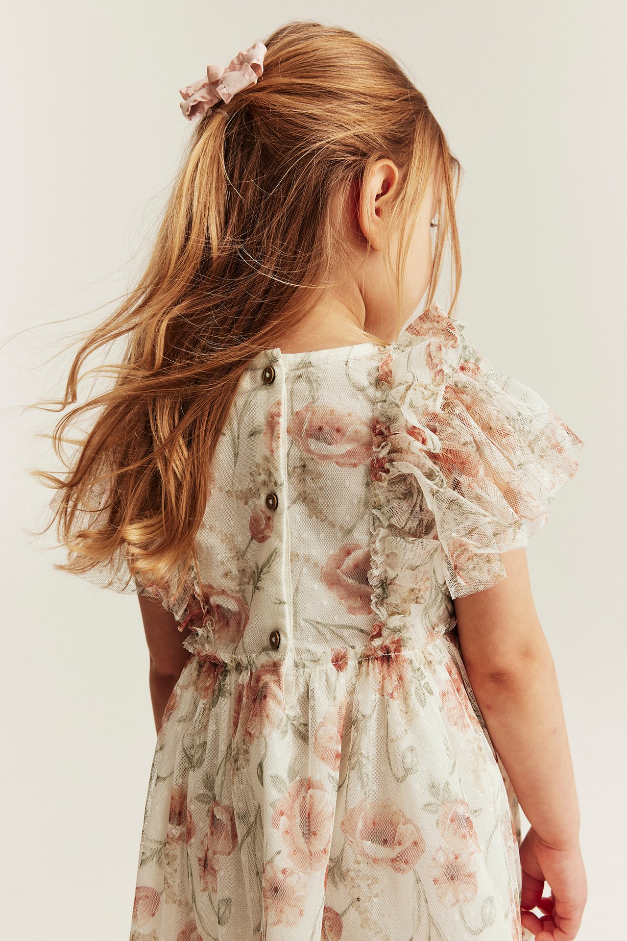 Barnets offwhite blommiga meshklänning, bakifrån, med volangärmar, knäppt rygg. Bär rosa hårtofs.