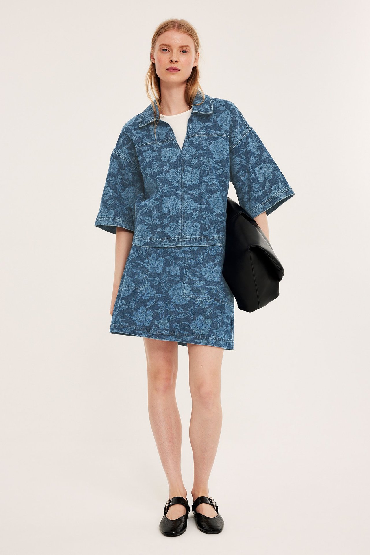 Kobieta w niebieskiej kwiecistej koszuli jeansowej, pasującej spódnicy, białym t-shircie, czarnych butach, z torbą.