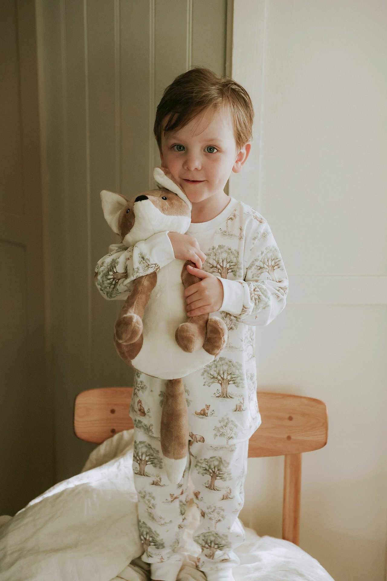 Lapsi edestä, yllään luonnonvalkoiset metsäkuvioiset puuvillapyjamat, pitäen pehmokettua.