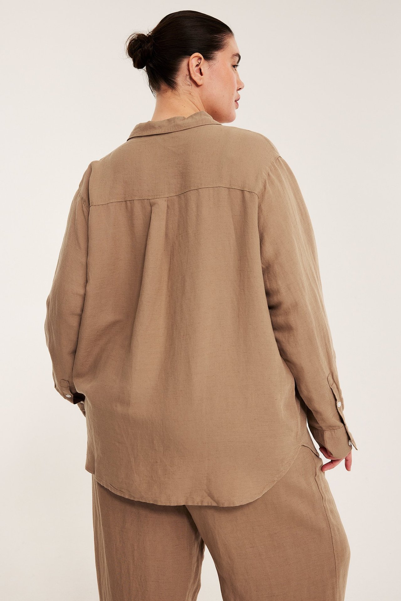 Ryggvy av en kvinna i en oversized beige linneskjorta med rundad fåll och matchande byxor.