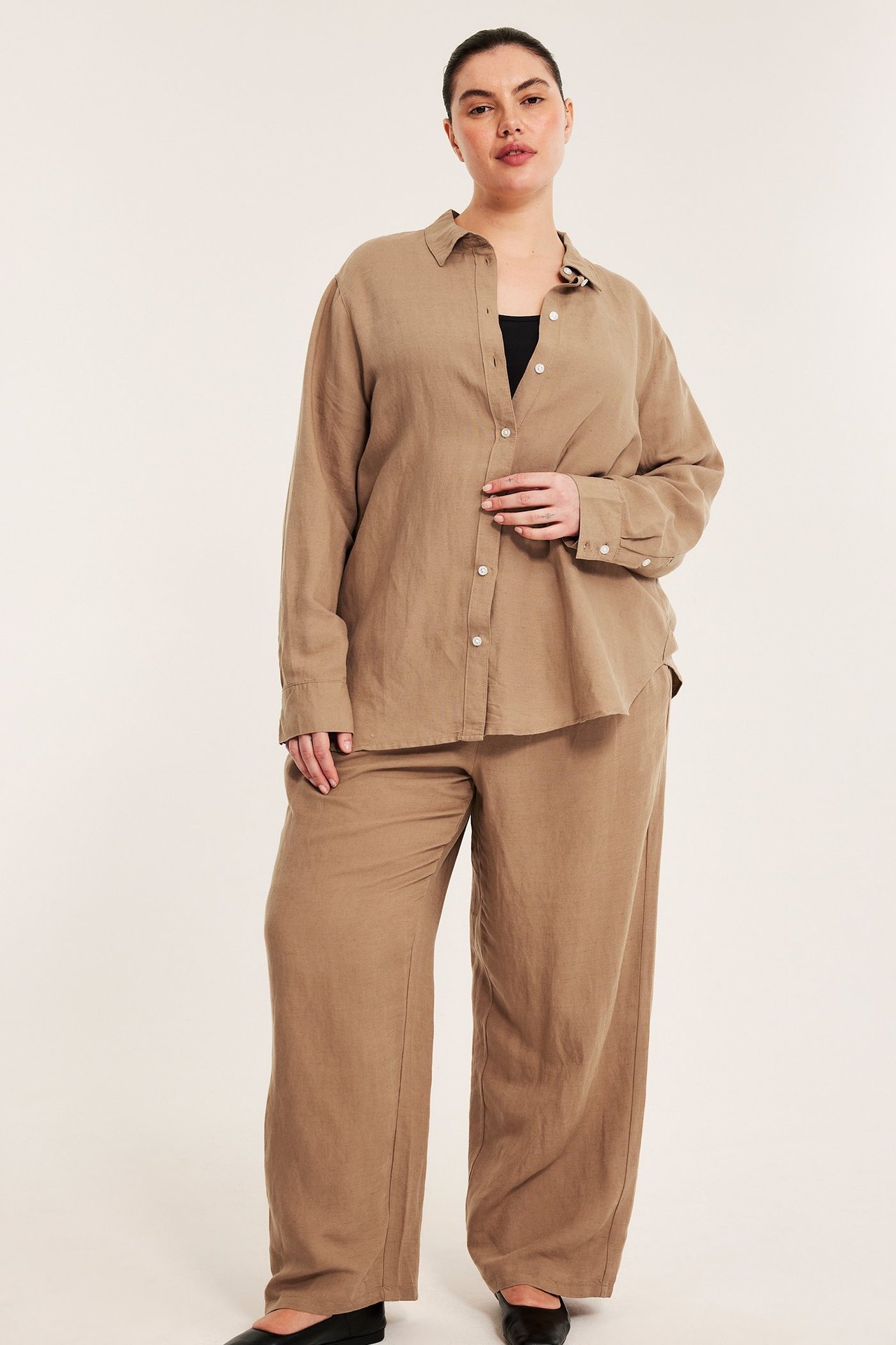 Front view: Kvinna i öppen beige linneblandningsskjorta över svart topp, matchande beiga byxor, svarta platta skor.