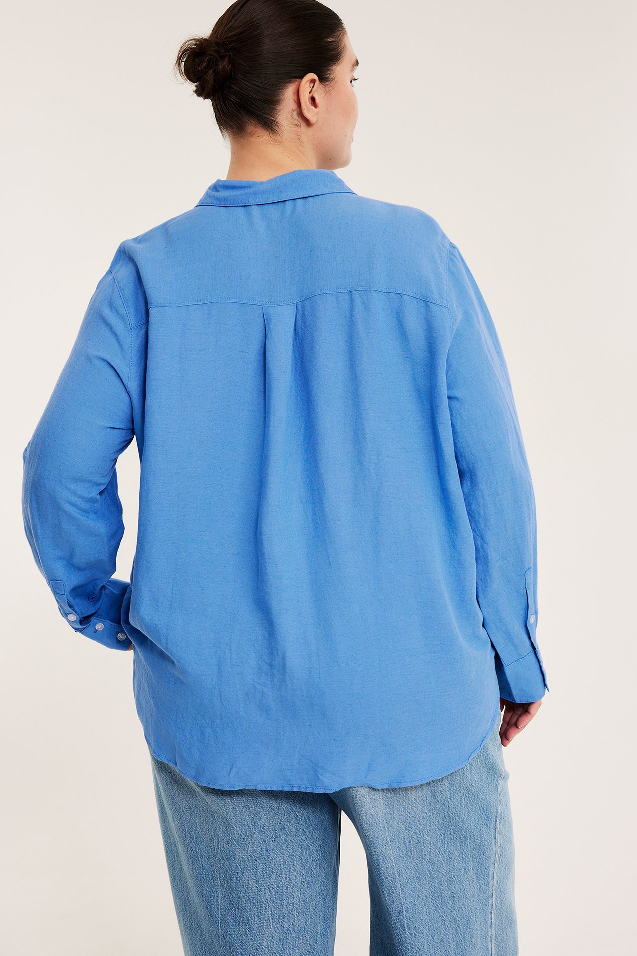 Bakifrån på en kvinna i en löst sittande blå linneblandningsskjorta och ljusblå jeans.