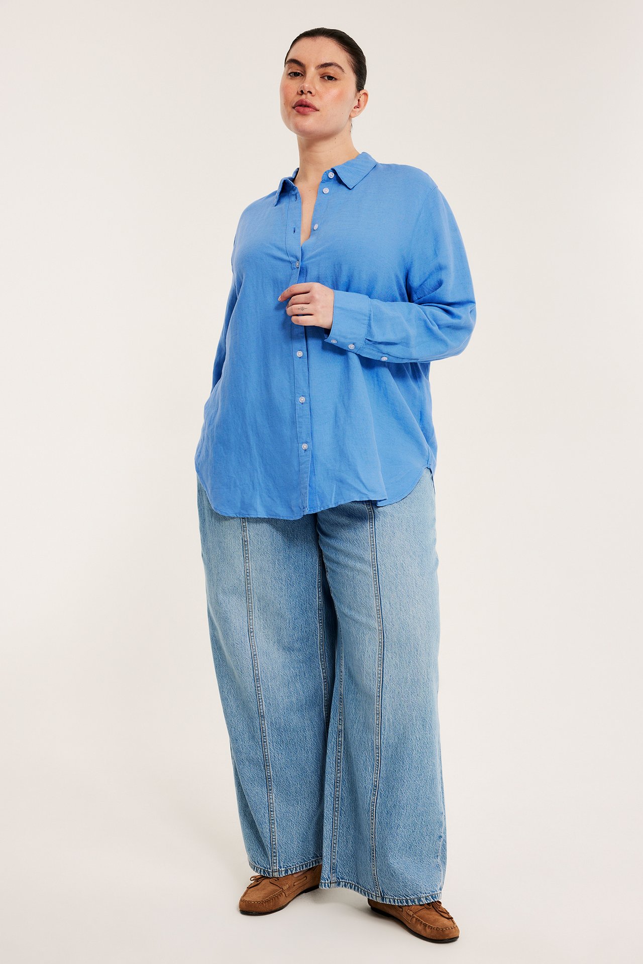 Framifrån: kvinna i lös blå linneblandad skjorta, ljusblå vida jeans och bruna mockasiner.