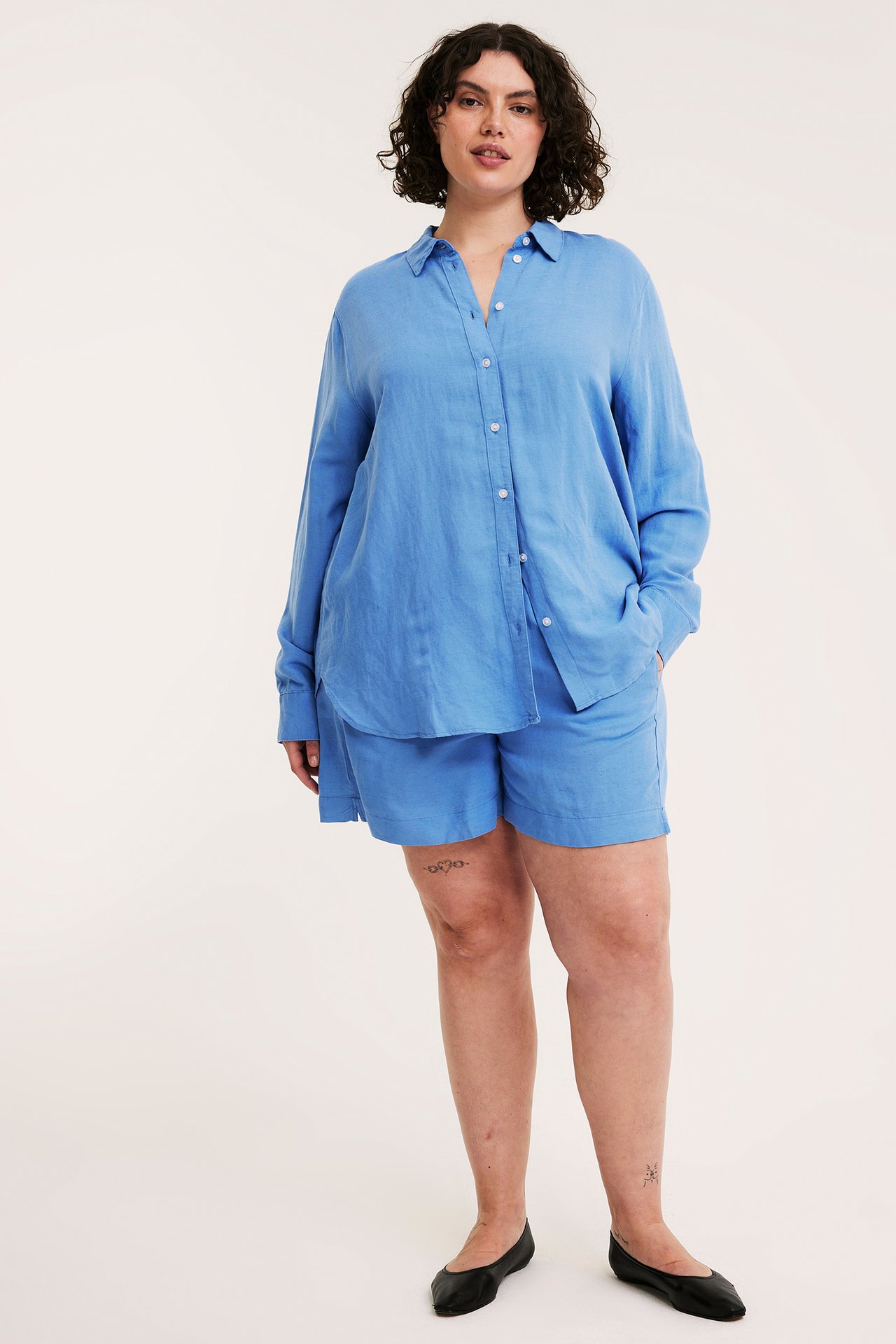 Kvinne forfra: blå linskjorte med hvite knapper, matchende blå shorts og svarte ballerinasko.