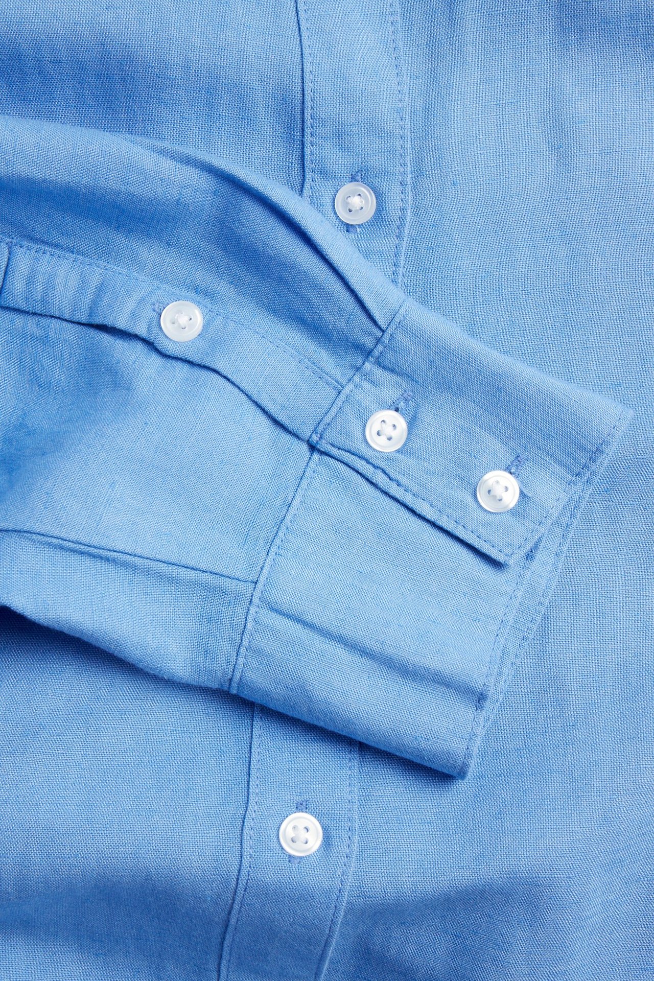 Zbliżenie niebieskiej koszuli z mieszanki lnu, pokazujące białe guziki na mankiecie i plisie.