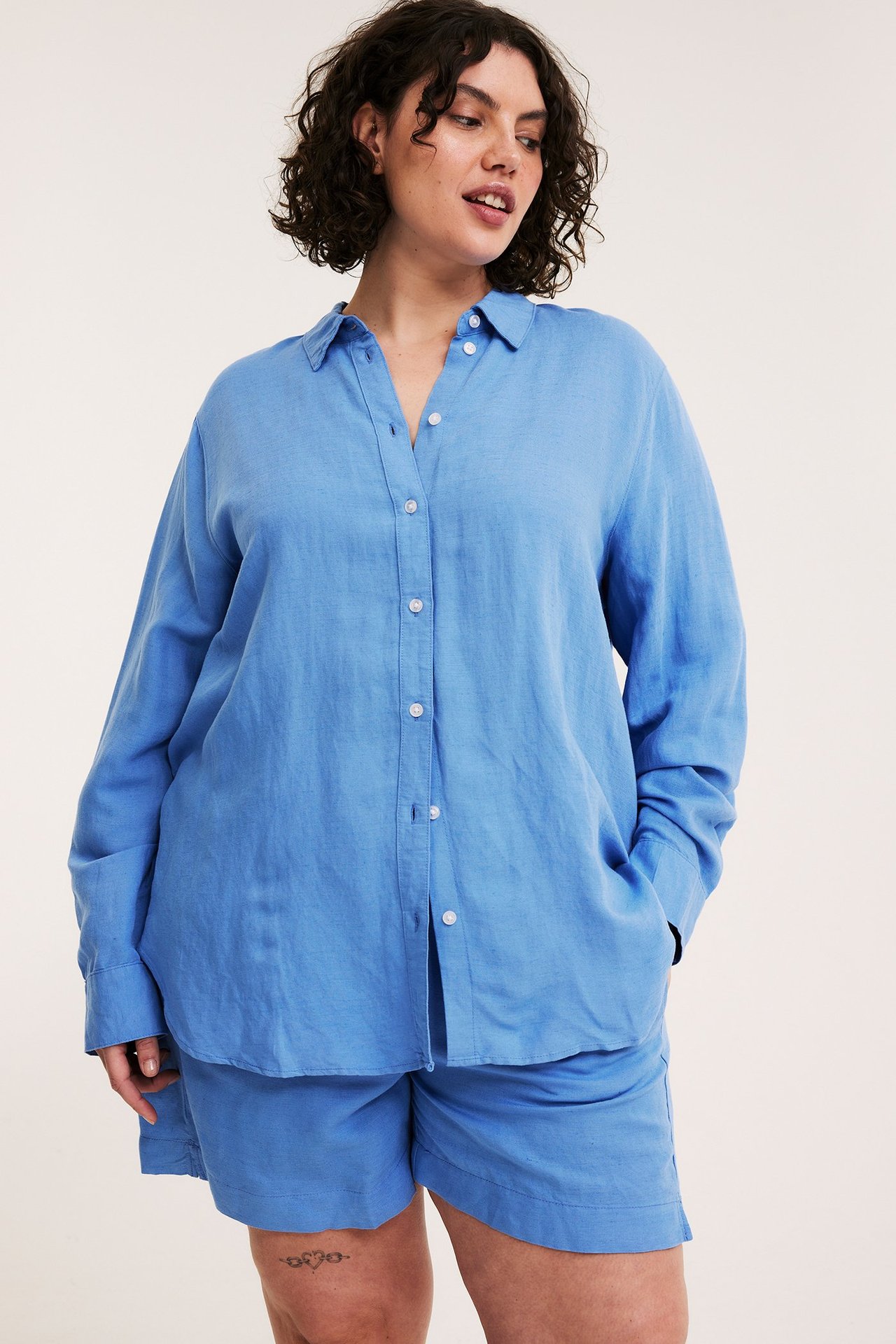 Widok z przodu: Kobieta w niebieskiej koszuli oversize z mieszanki lnu, z białymi guzikami i pasującymi niebieskimi szortami.