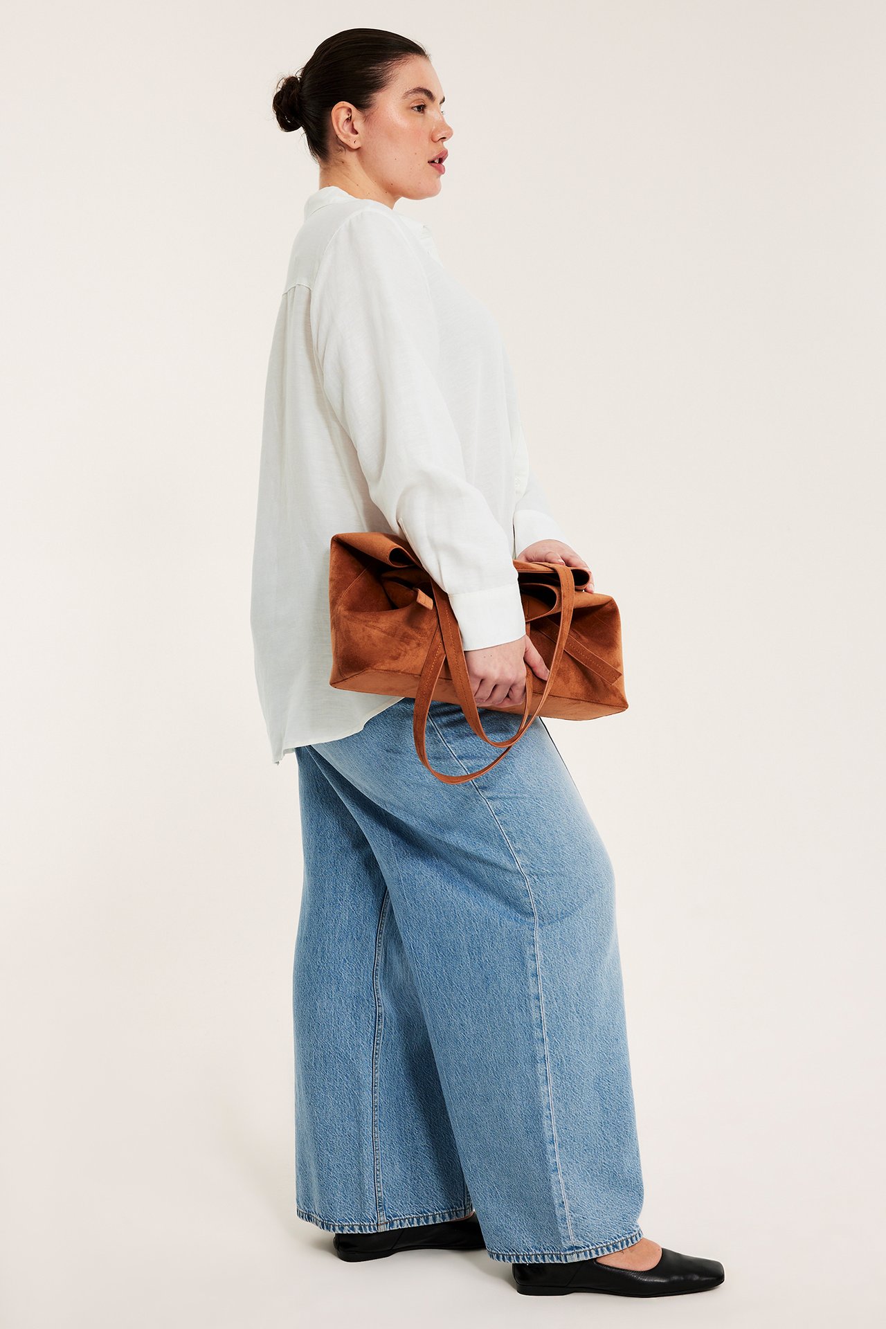 Sidovy av en kvinna i vit linneskjorta, ljusblå vida jeans, svarta platta skor, håller brun handväska.