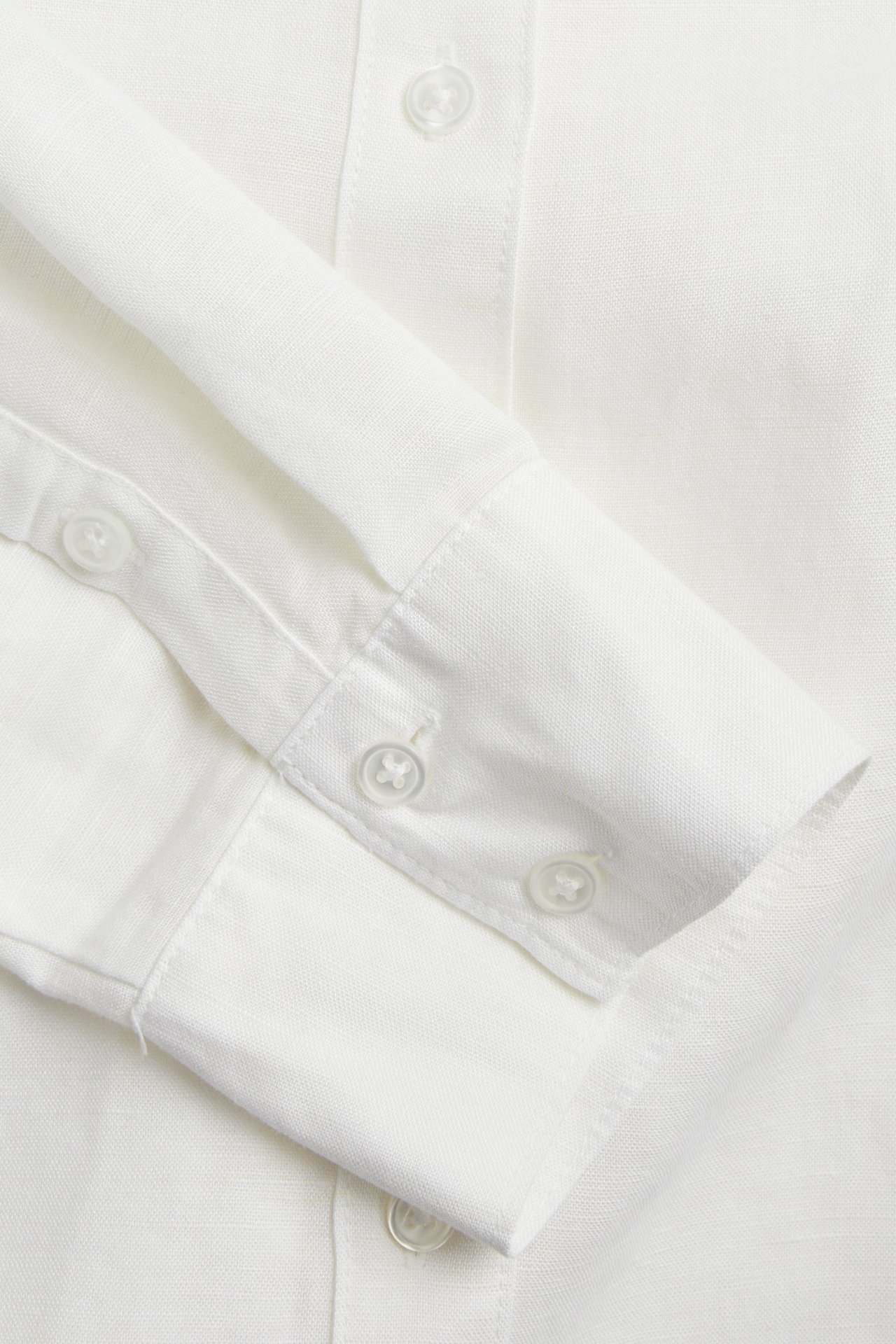 Zbliżenie białej koszuli z mieszanki lnu, ukazujące guziki na plisie i mankiecie.