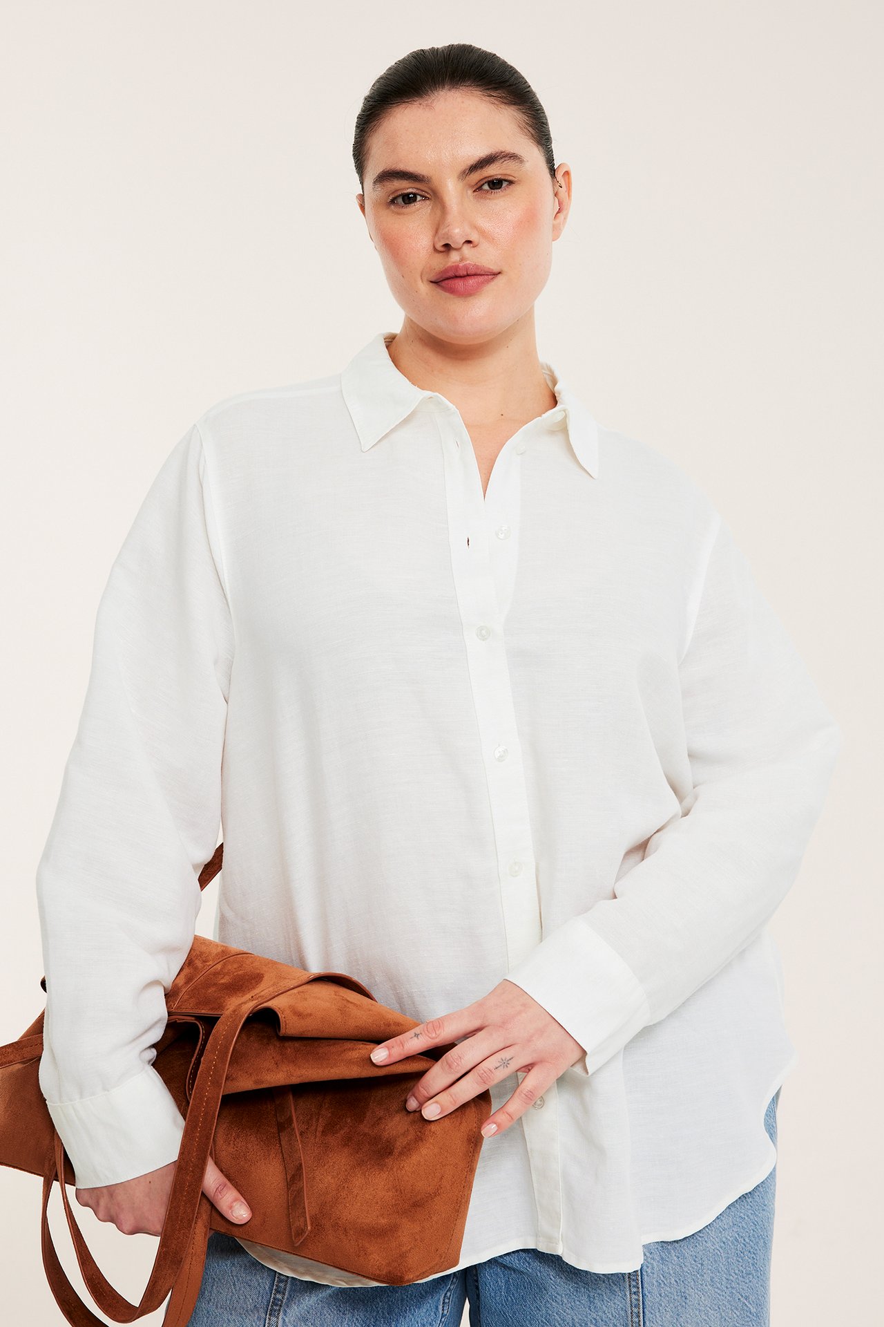 Frontbild av kvinna i vit linneblandningsskjorta, blå jeans, och som håller en brun mockaväska.