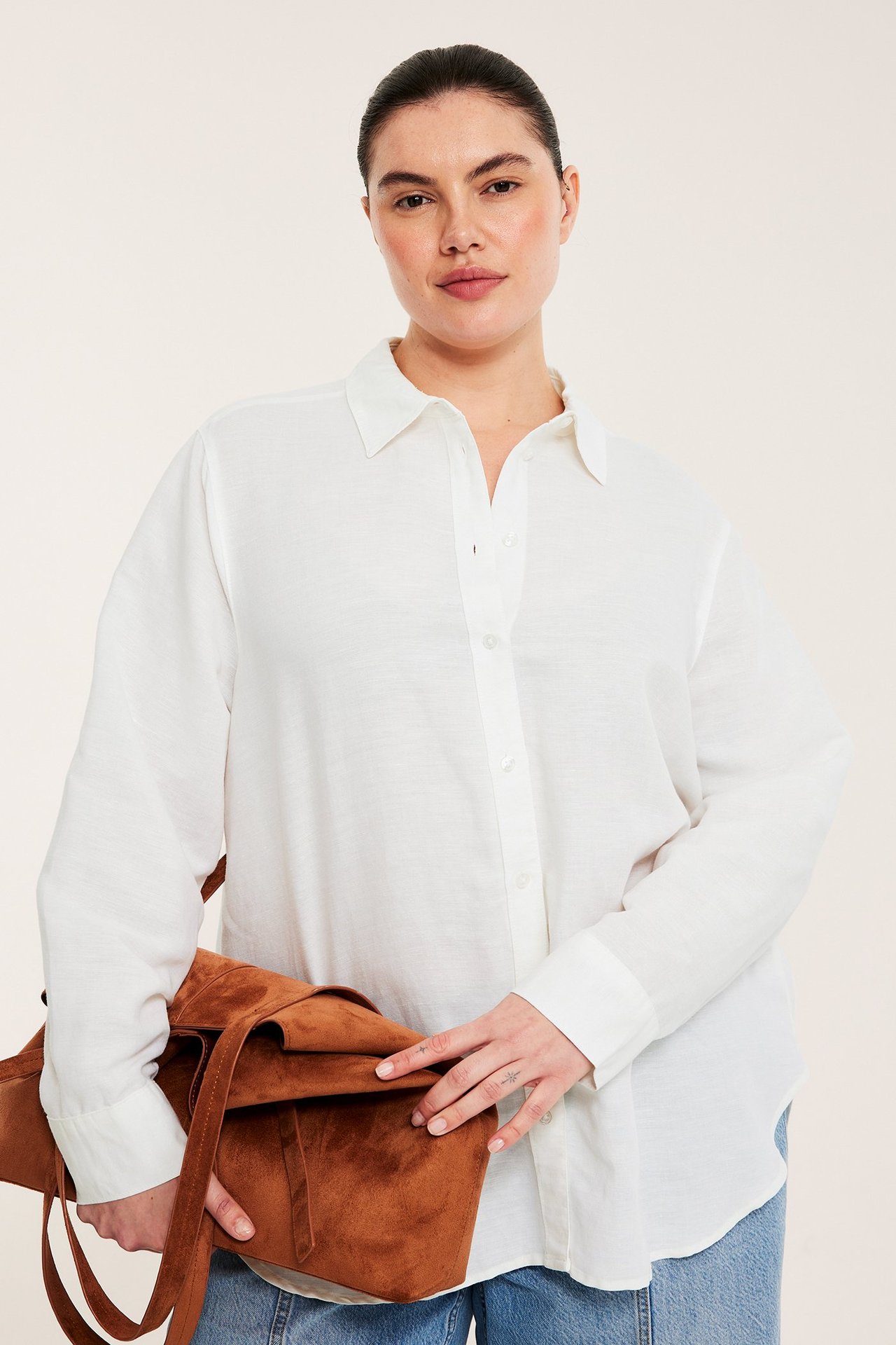 Frontbild av kvinna i vit linneblandningsskjorta, blå jeans, och som håller en brun mockaväska.