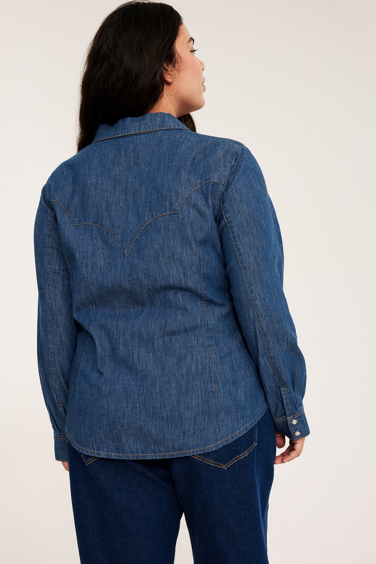 Bak-høyre visning av kvinne i tettsittende blå denimskjorte med western-ok og lyse knapper, over mørke jeans.