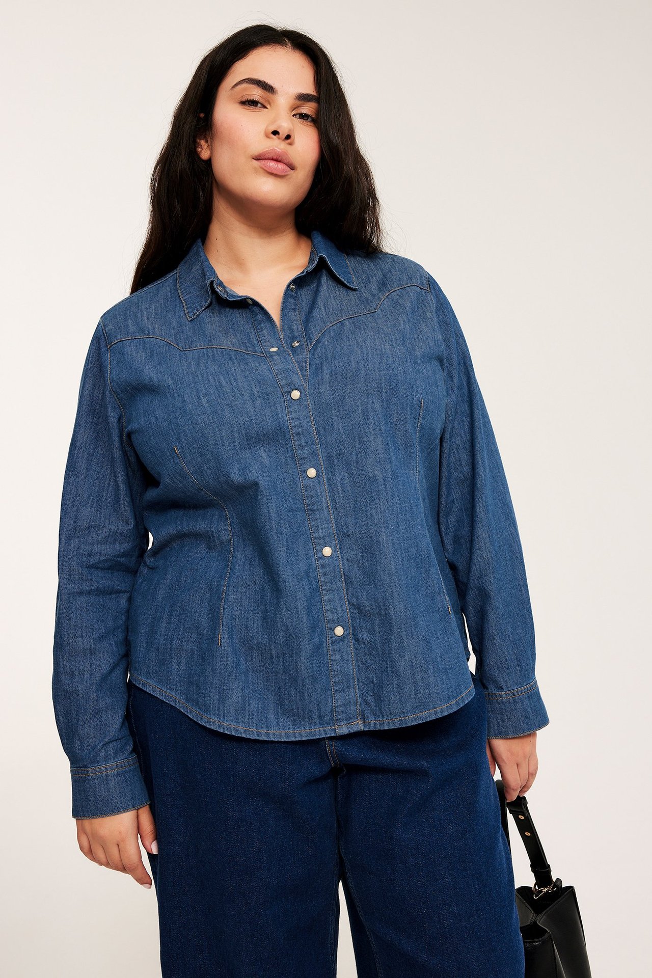 Forfra: Kvinne i blå denimskjorte med hvite trykknapper, mørkeblå jeans, holder svart håndveske.