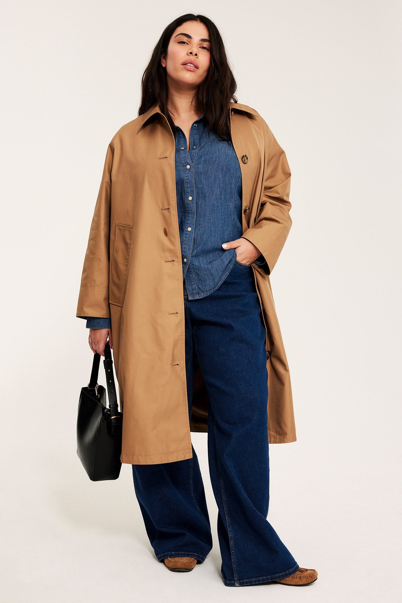 Kvinna bär figursydd blå jeansskjorta med knappar, öppen kamelfärgad trenchcoat, vida mörka jeans, svart handväska.