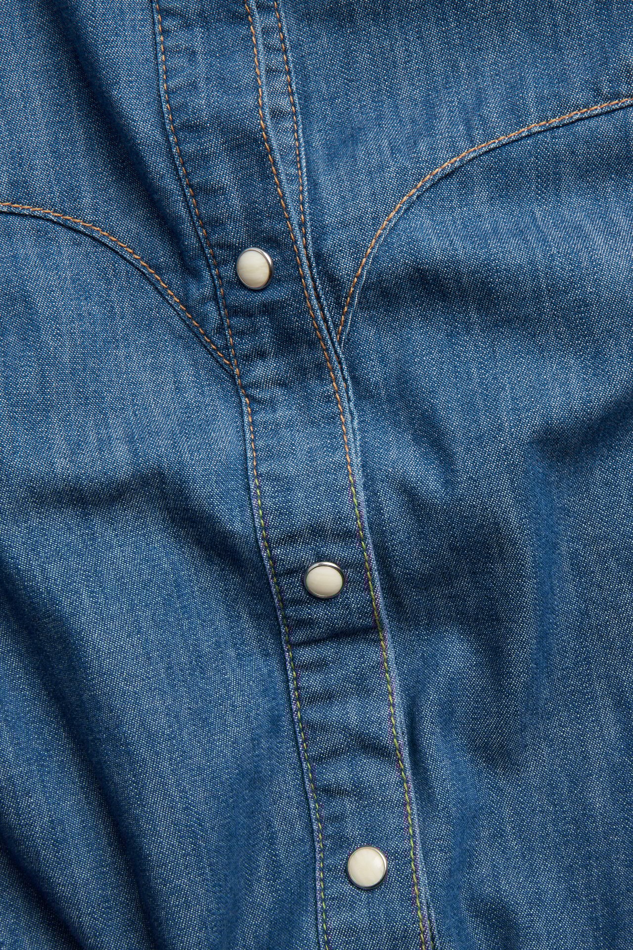 Lähikuva edestä sinisestä farkkupaidasta, jossa on western-kaarroke ja luonnonvalkoiset painonapit.