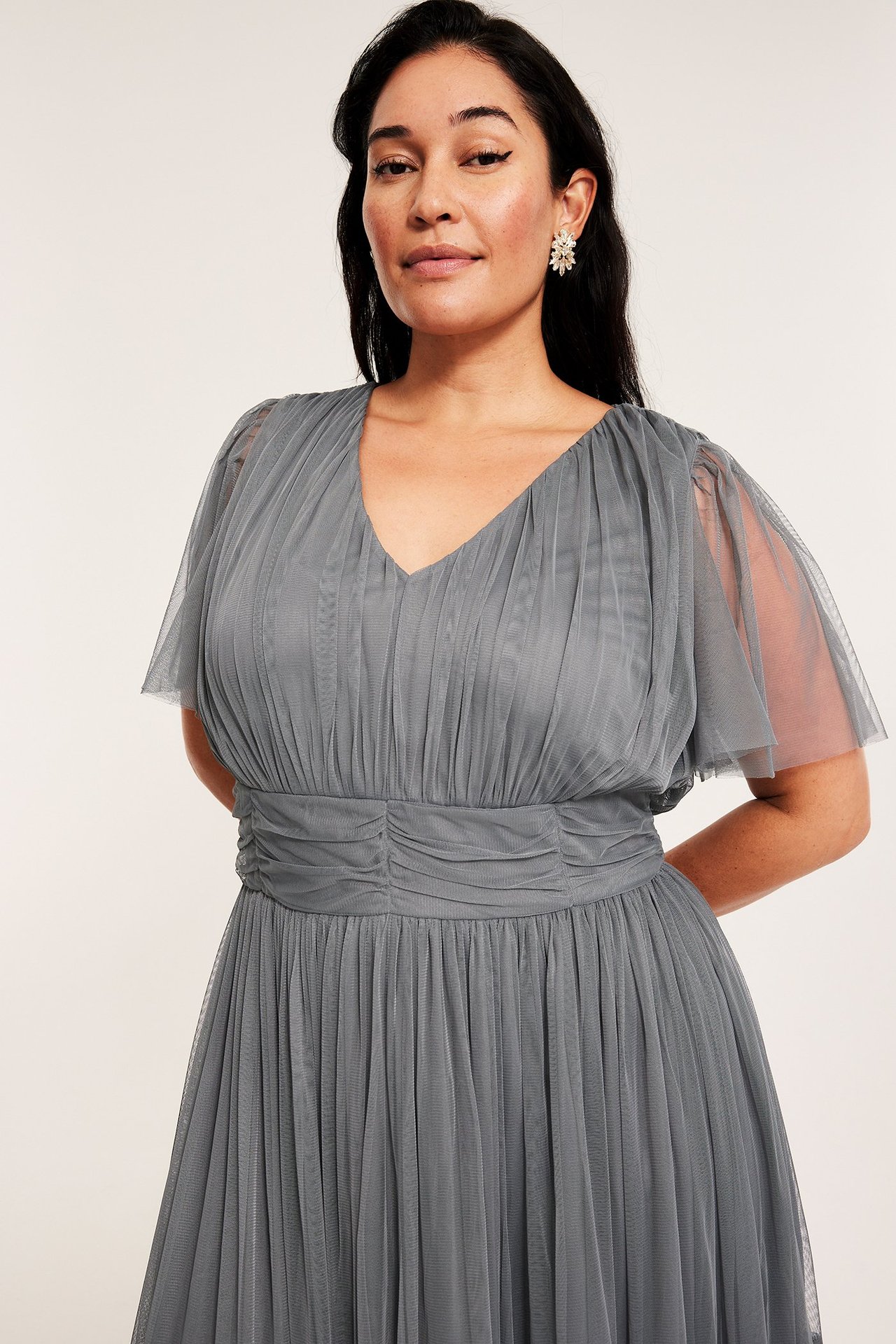 Edestä: nainen harmaassa tyllimekossa, V-pääntie, rypytetty yläosa, leijailevat hihat ja korvakorut.