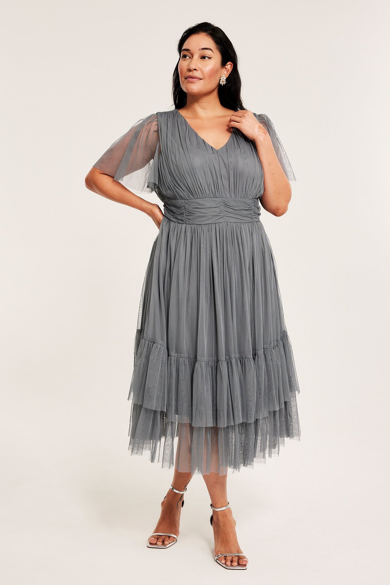 Edestä: nainen yllään harmaa V-aukkoinen tyllimekko, jossa rypytetty vyötärö, röyhelöhame ja hopeasandaalit.