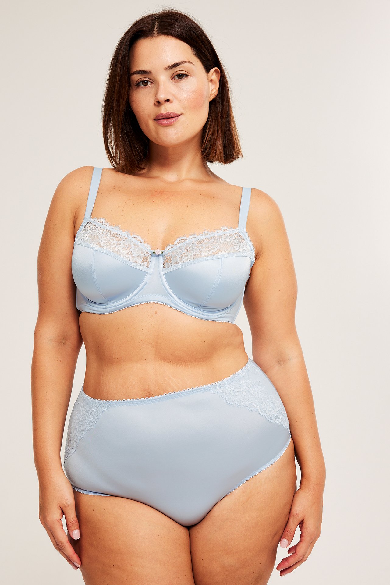 Edestä päin, nainen yllään vaaleansiniset korkeavyötäröiset pikkuhousut pitsiyksityiskohdilla ja yhteensopivat rintaliivit.