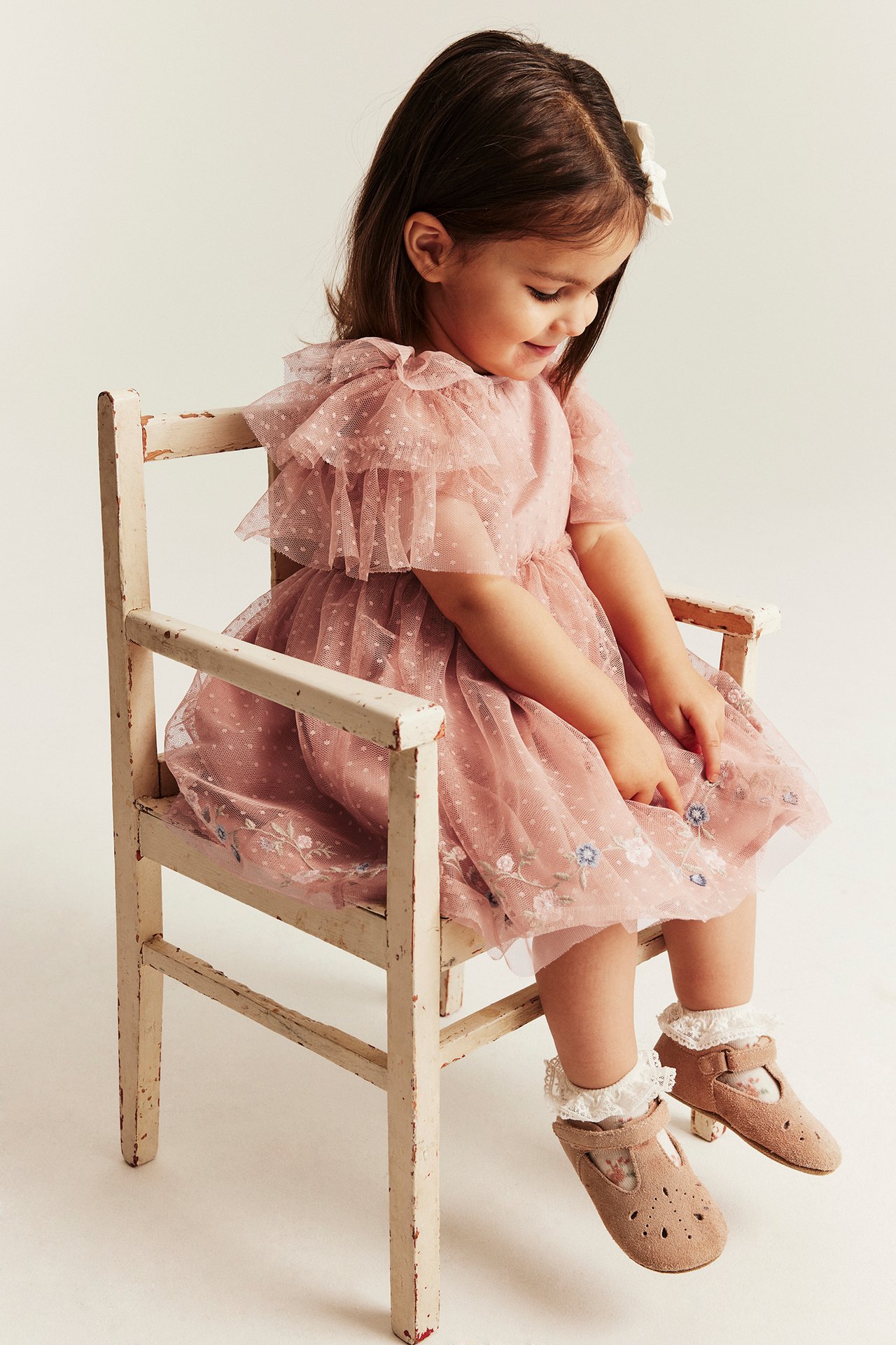 Barn i rosa prikkete meshkjole med volangermer, blomsterbroderi, hvite blondestrømper og beige sko.