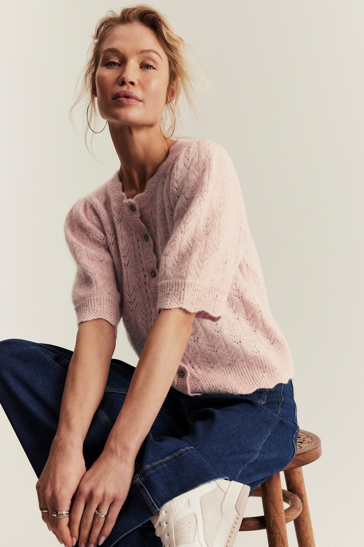 Kobieta w różowym wełnianym kardiganie z krótkim rękawem, niebieskich jeansach i złotych kolczykach.