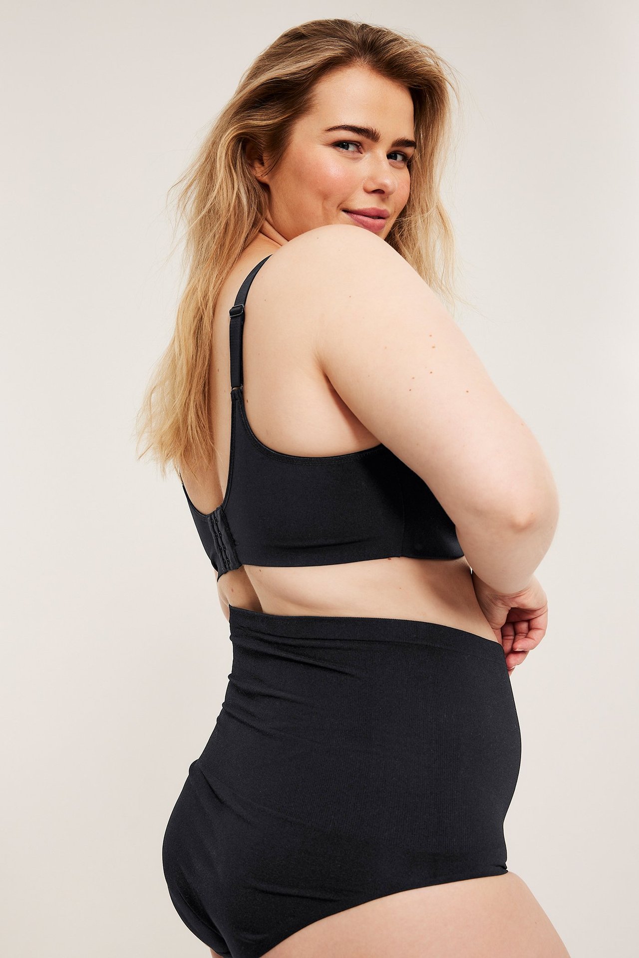 Sivukuva: nainen mustissa saumattomissa rintaliiveissä takakiinnityksellä ja mustissa korkeissa alushousuissa.