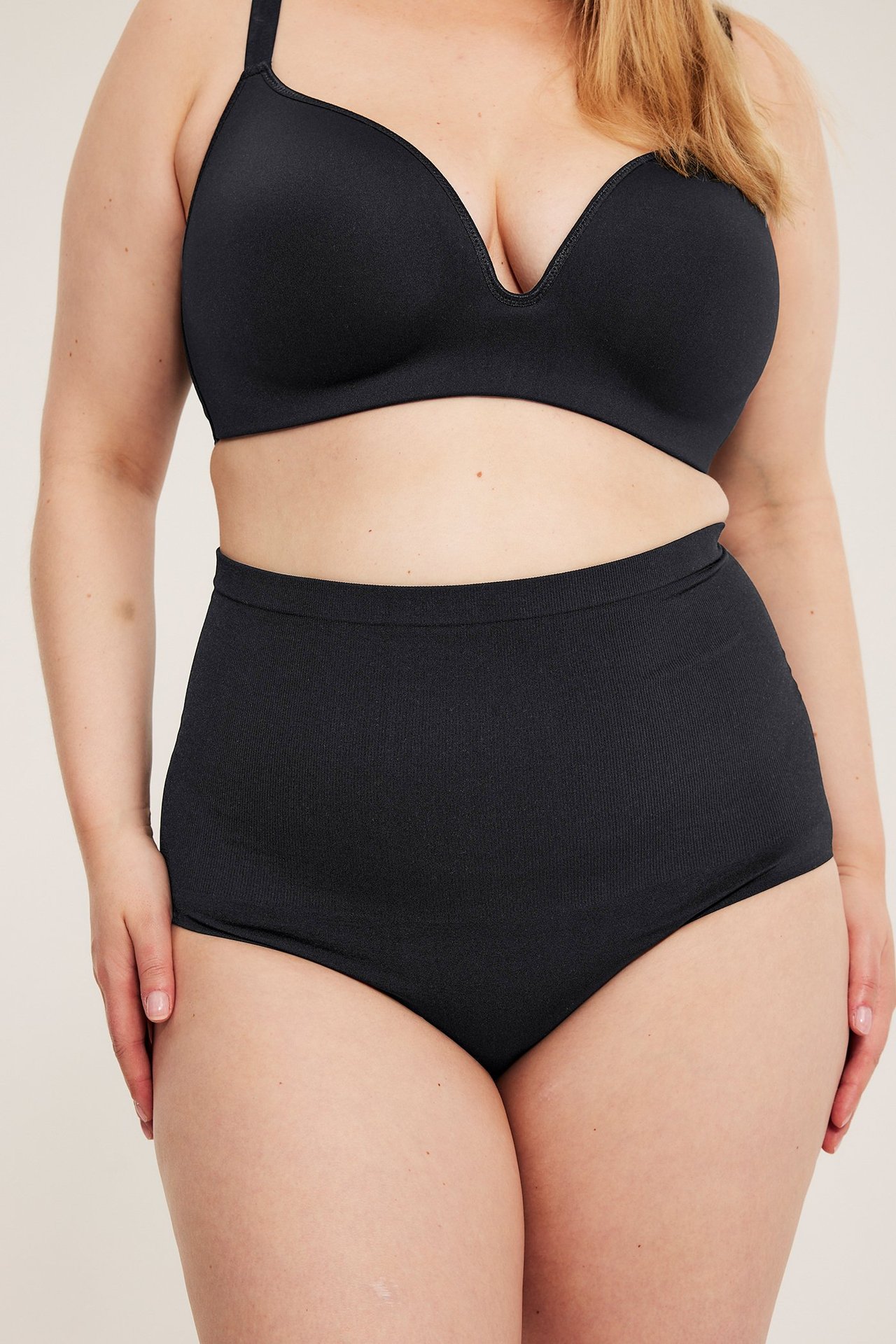 Edestä, nainen mustassa saumattomassa rintaliivissä ja korkeavyötäröisissä pikkuhousuissa.