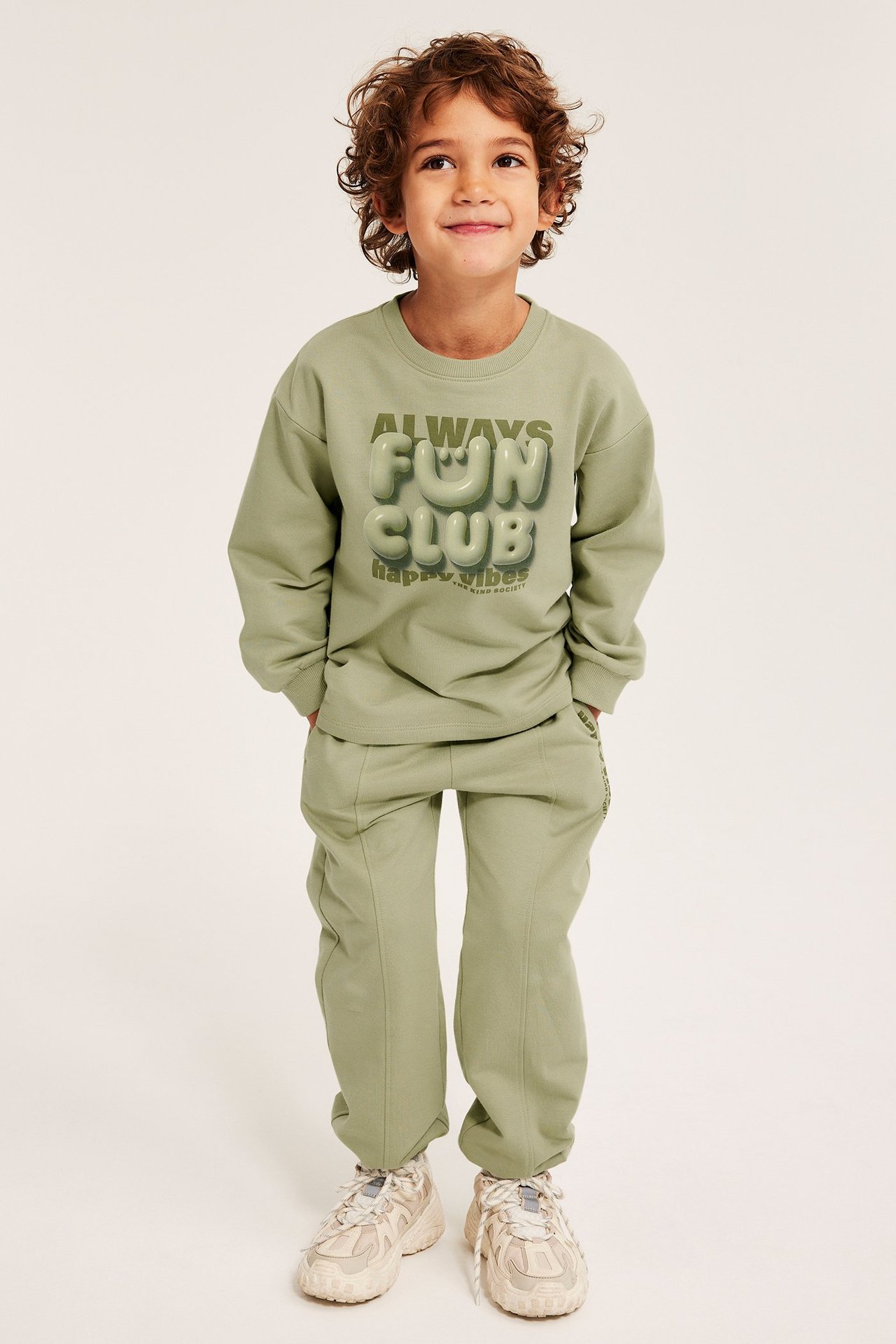 Widok z przodu: Dziecko w ciemnozielonych bawełnianych dresach z mankietami, zielonej bluzie z nadrukiem i beżowych trampkach.