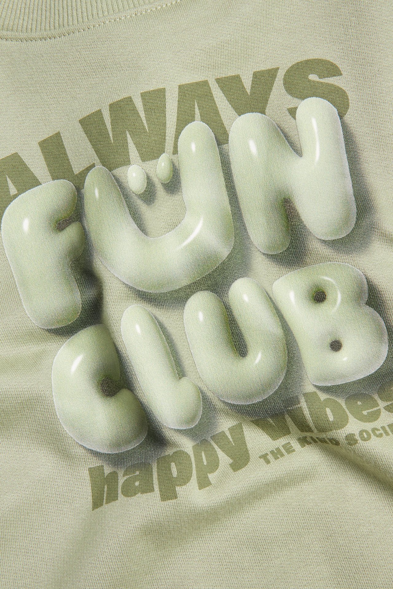 Zbliżenie na ciemnozieloną bawełnianą bluzę z wypukłym, błyszczącym nadrukiem "FUN CLUB" z przodu.