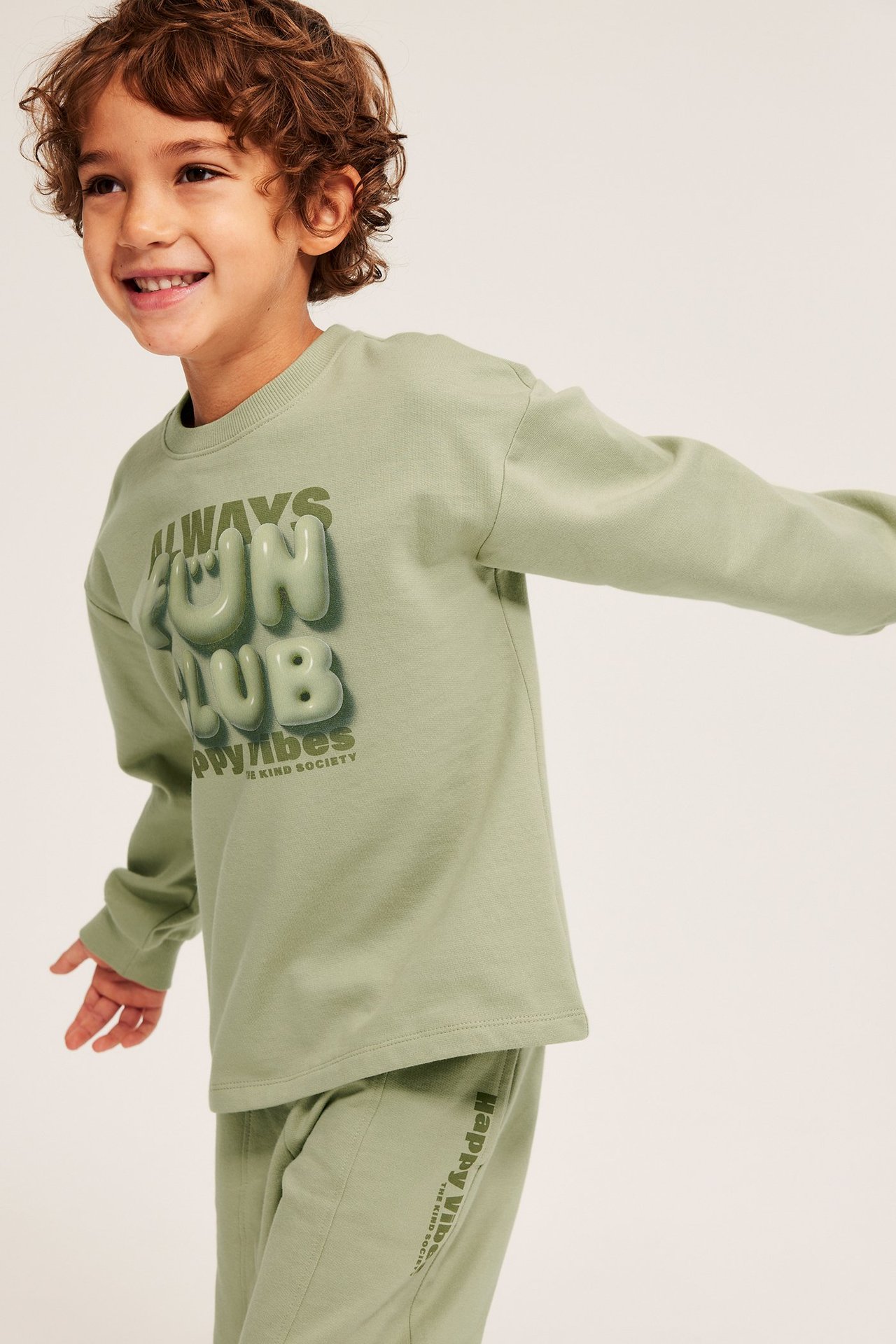 Edestäpäin: lapsi vaaleanvihreässä puuvillapaidassa 'ALWAYS FUN CLUB' -painatuksella ja housuissa.
