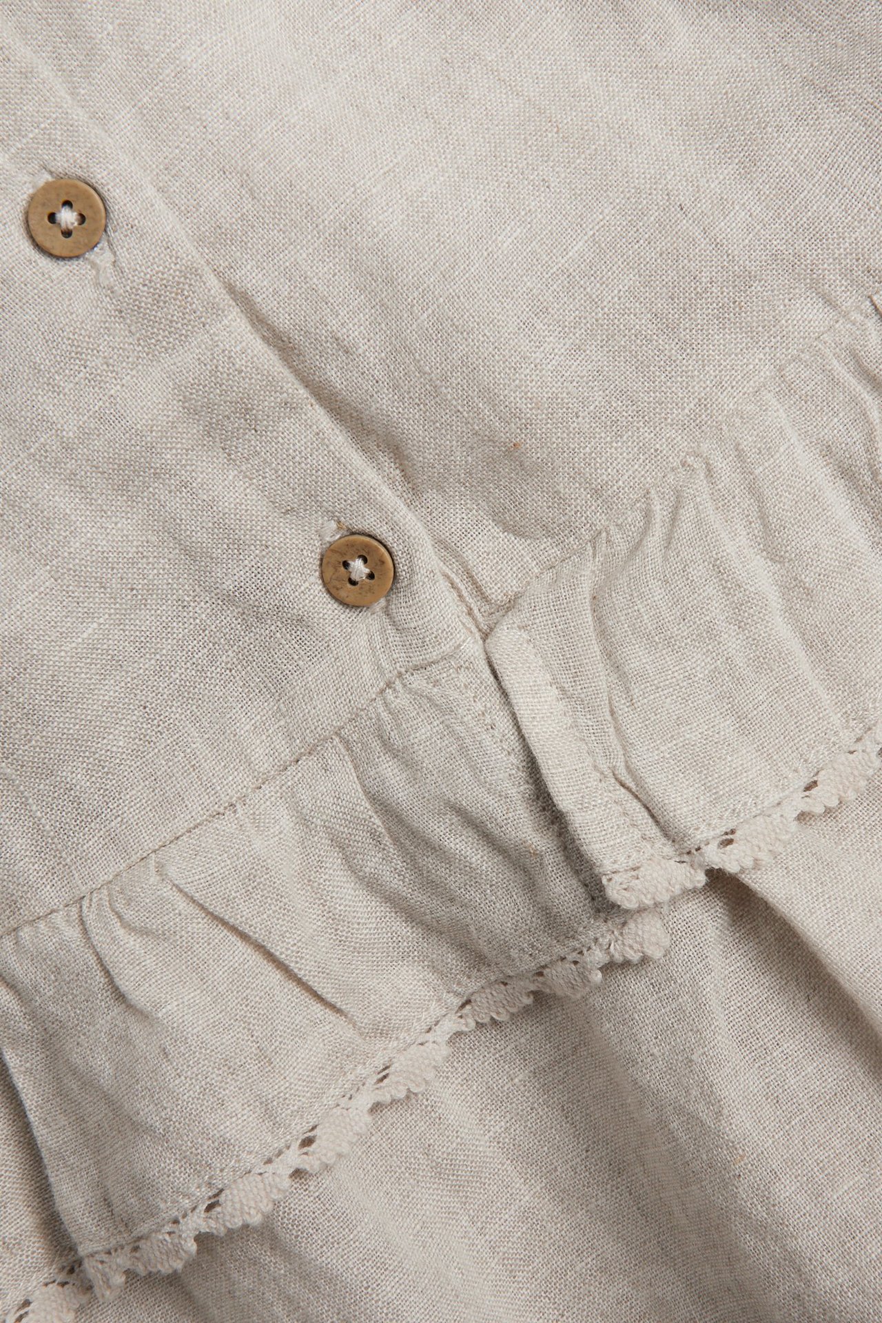 Zbliżenie jasnobeżowej lnianej sukienki z falbanami, koronkową lamówką i dwoma brązowymi drewnianymi guzikami.