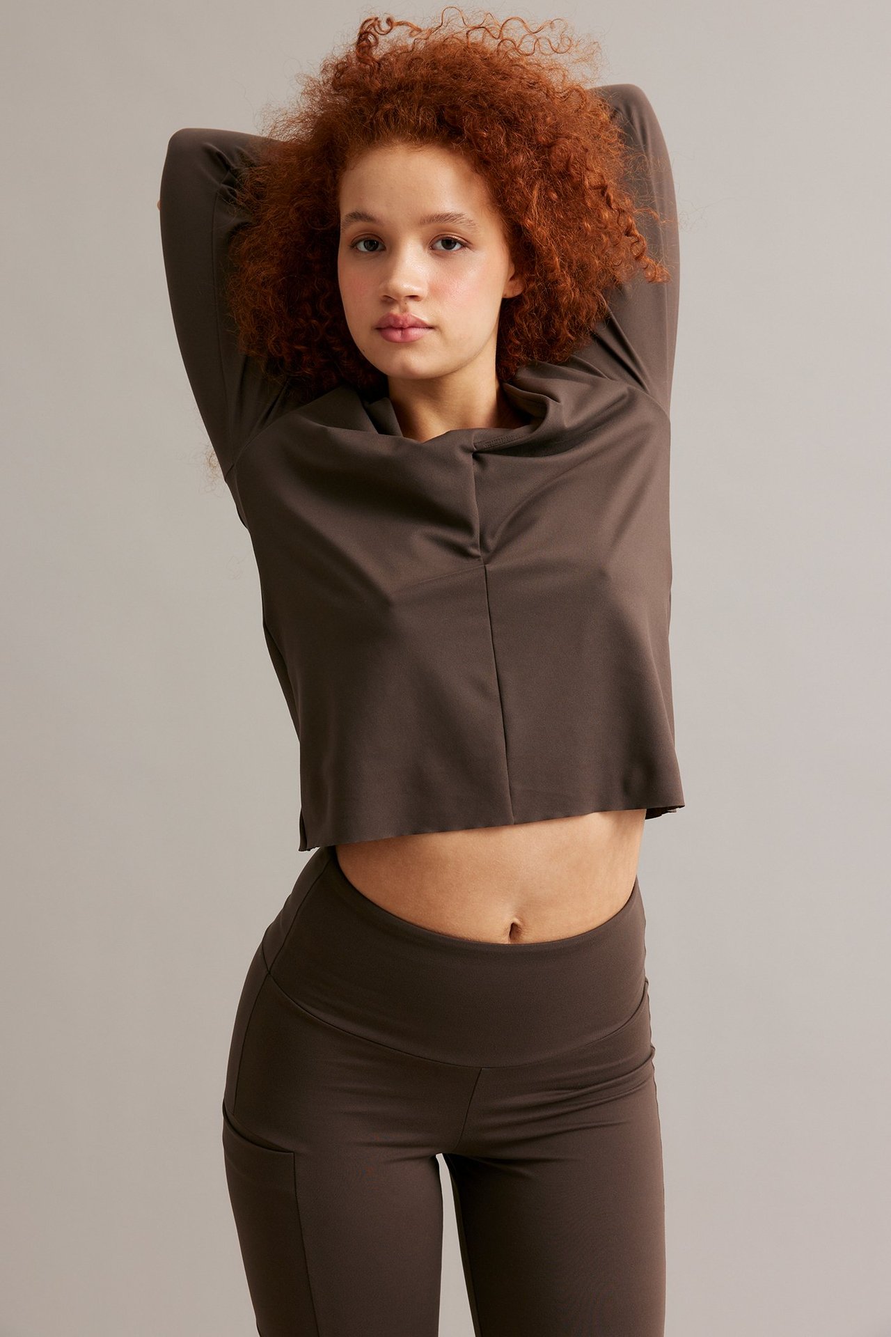 Frontvisning av en kvinne i brun, kort V-hals treningstopp med omslagsdetalj og matchende brune leggings.
