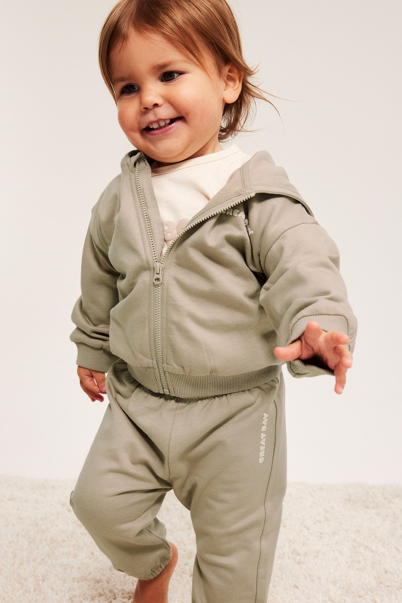 Forfra, baby i beige bomullsmykisbukser med trykk og matchende hettegenser med glidelås.