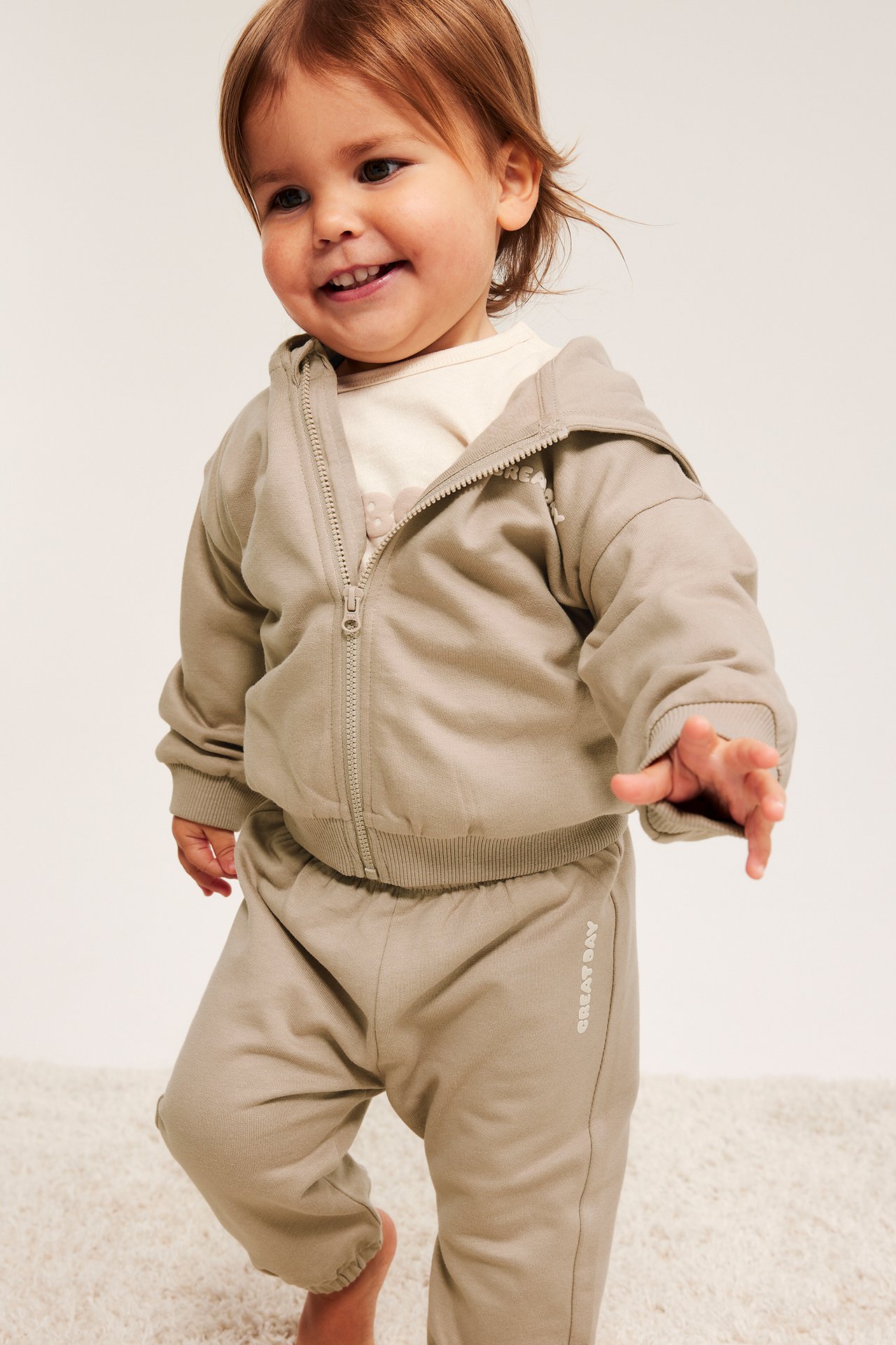 Baby i beige bomullsjoggebukse med sidetrykk, matchende hettegenser med glidelås og lys topp.
