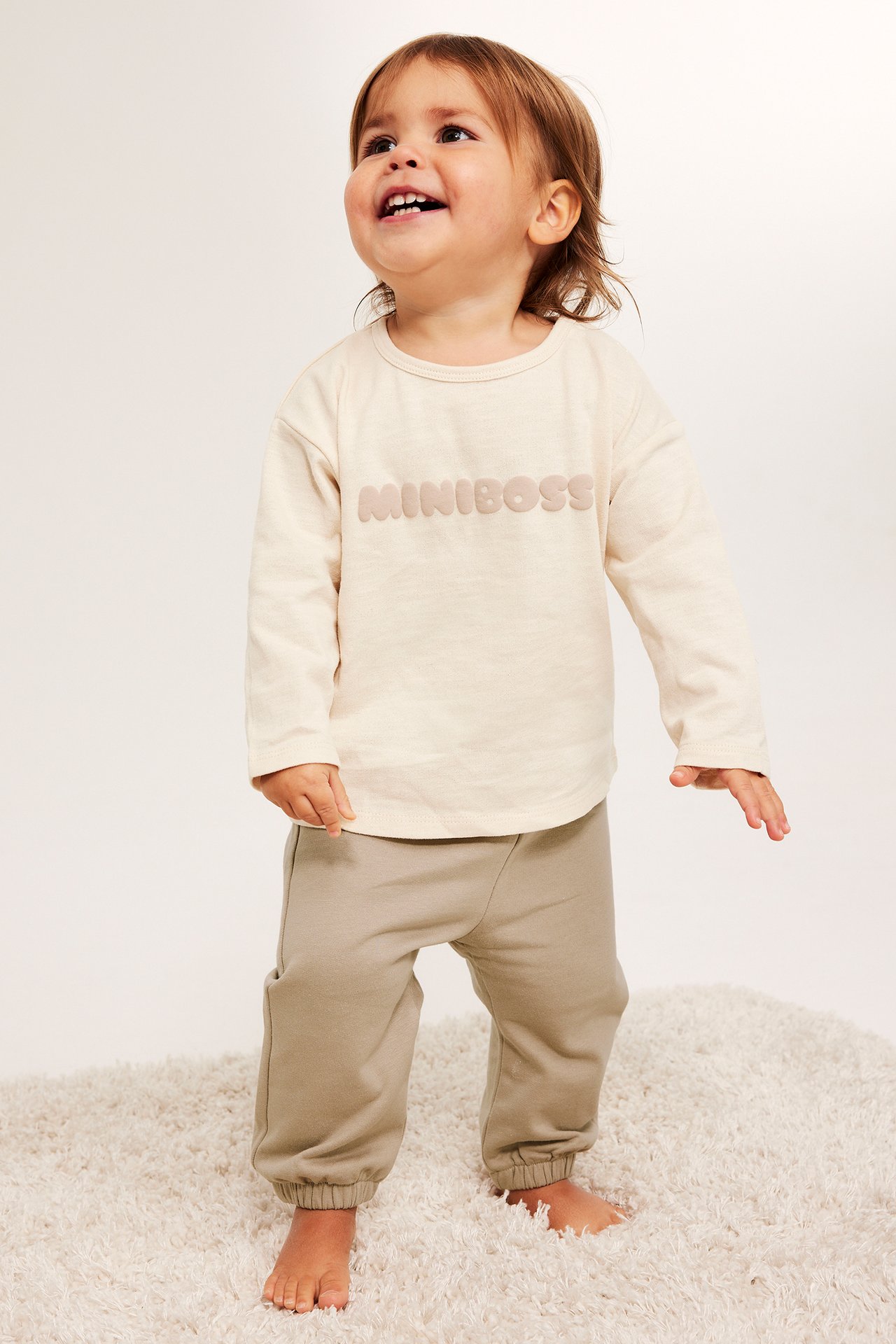 Frontvisning av en baby i løse beige joggebukser med ribbekanter og en kremfarget 'MINIBOSS' langermet topp.