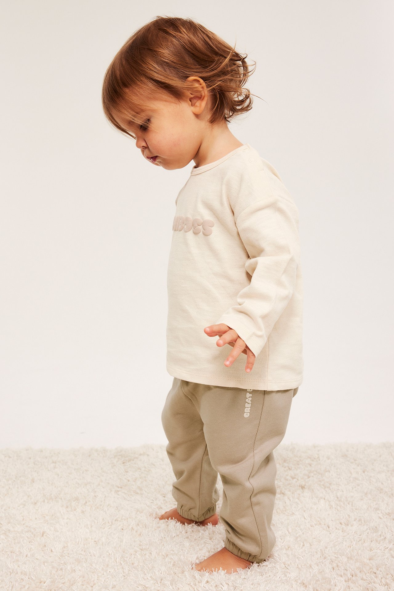 Widok z boku niemowlęcia w beżowych bawełnianych spodniach dresowych z bocznym nadrukiem i haftowanej kremowej bluzce.
