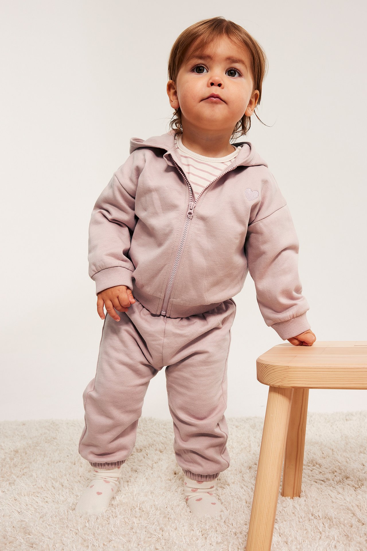 Widok z przodu: niemowlę w różowych bawełnianych dresach i pasującej bluzie z kapturem na zamek z haftowanym sercem.