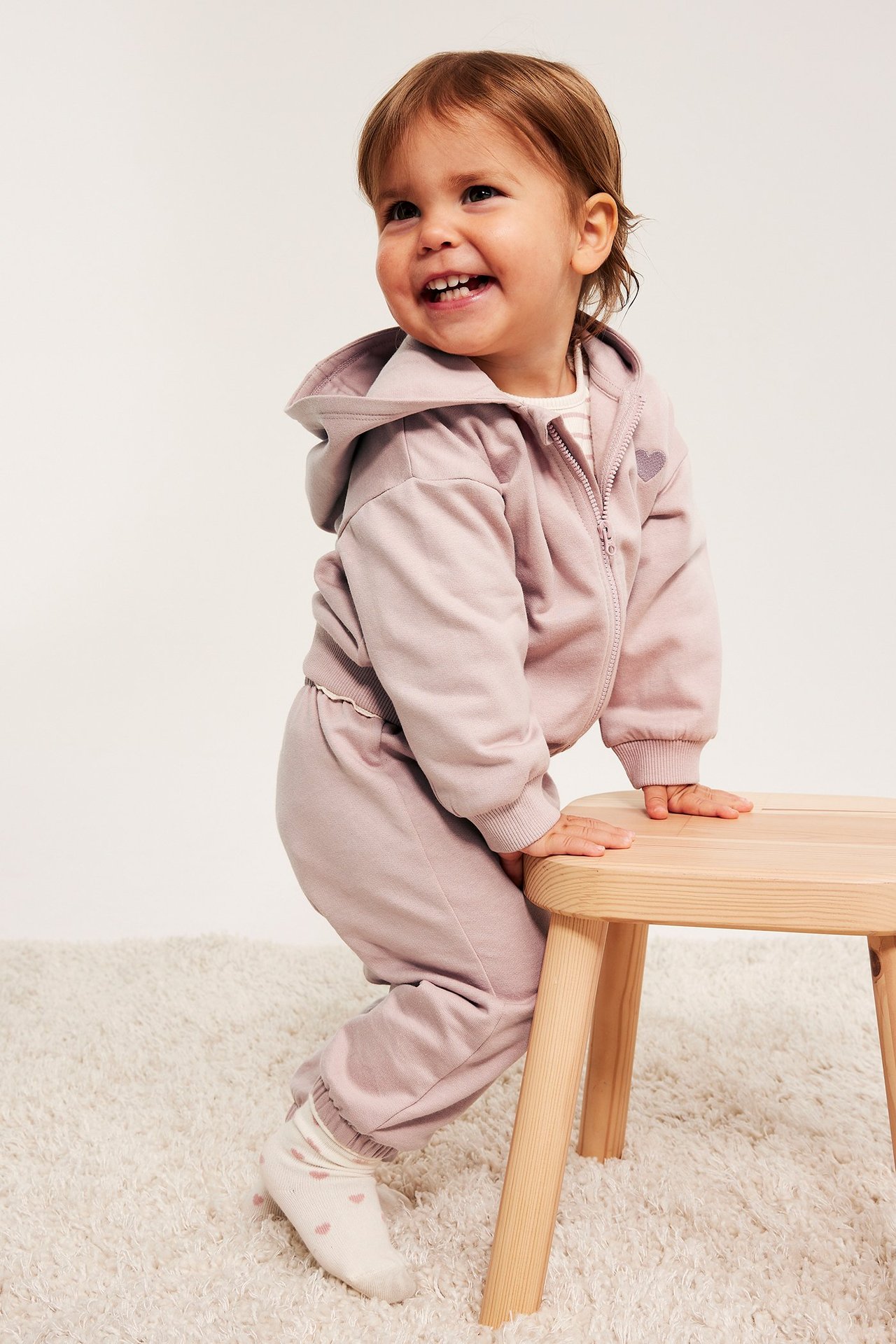 Uśmiechnięte dziecko z przodu w jasnoróżowym bawełnianym dresie z kapturem, haftem w serce i białych skarpetkach.