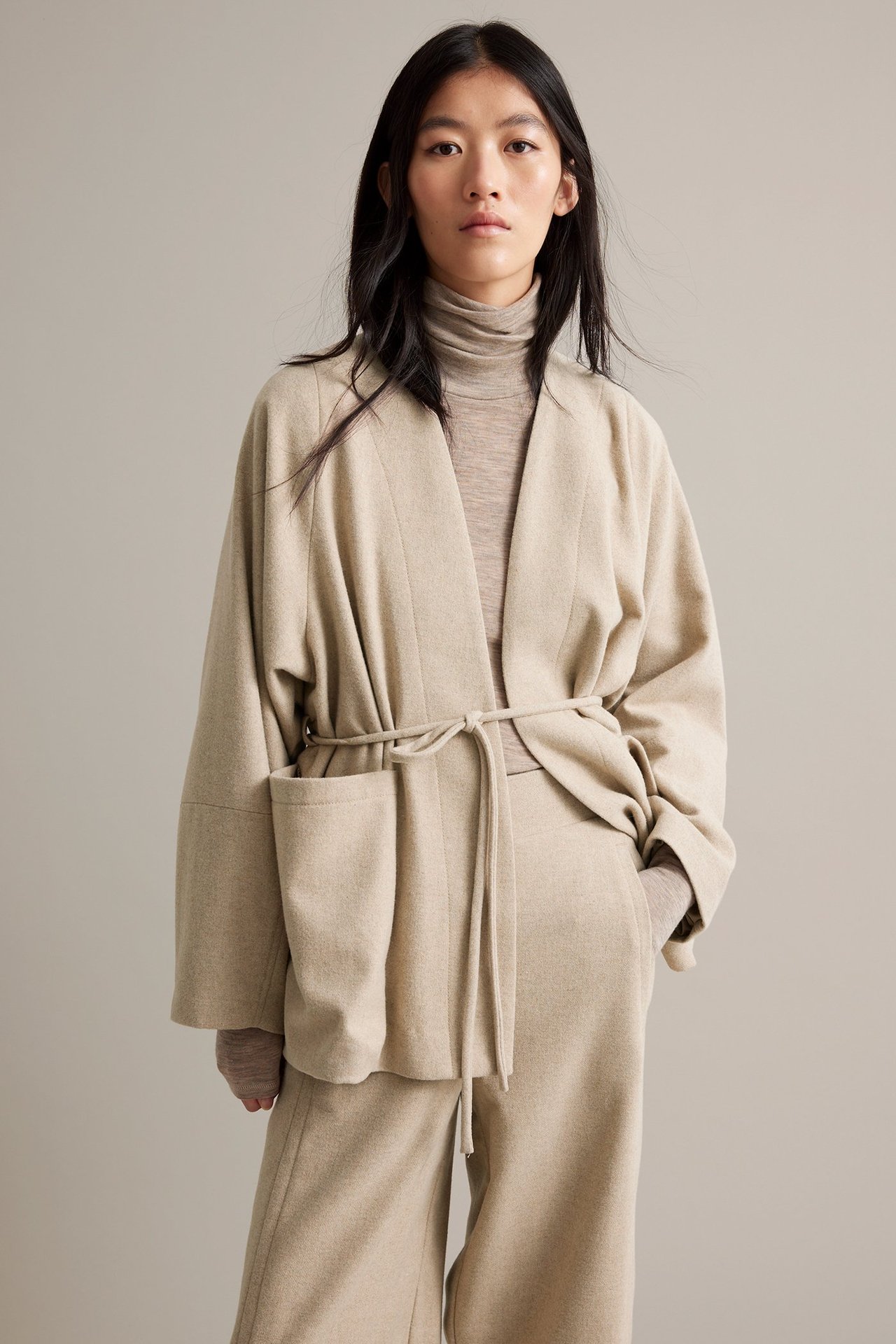 Nainen beigeä melsiroitua villa-sekoitejackkia, vyöllä ja rullakauluksella.