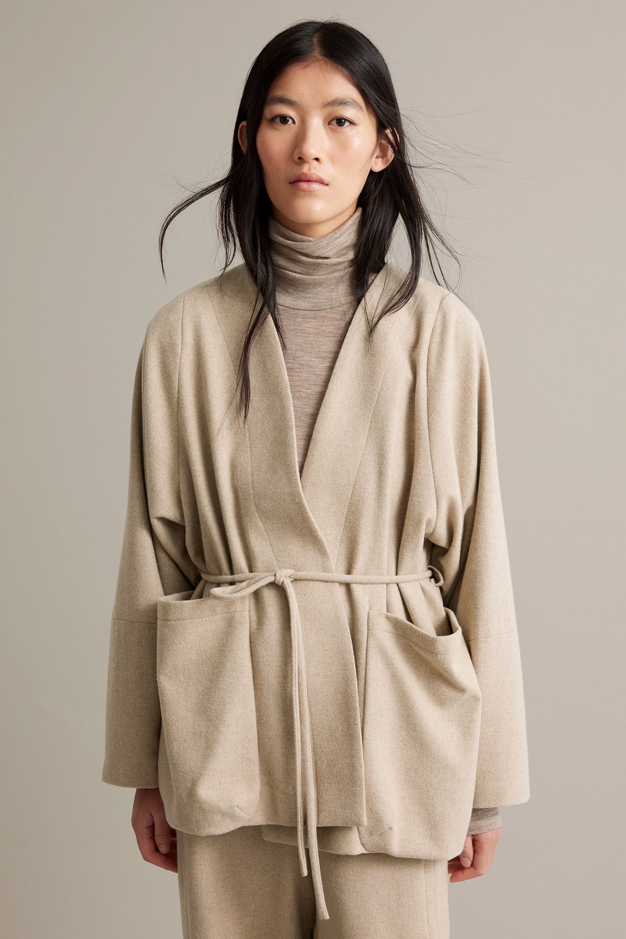 Nainen beigessä melangelaatikkovillakangas takki, vuorattu, vyö, etunäkymä.