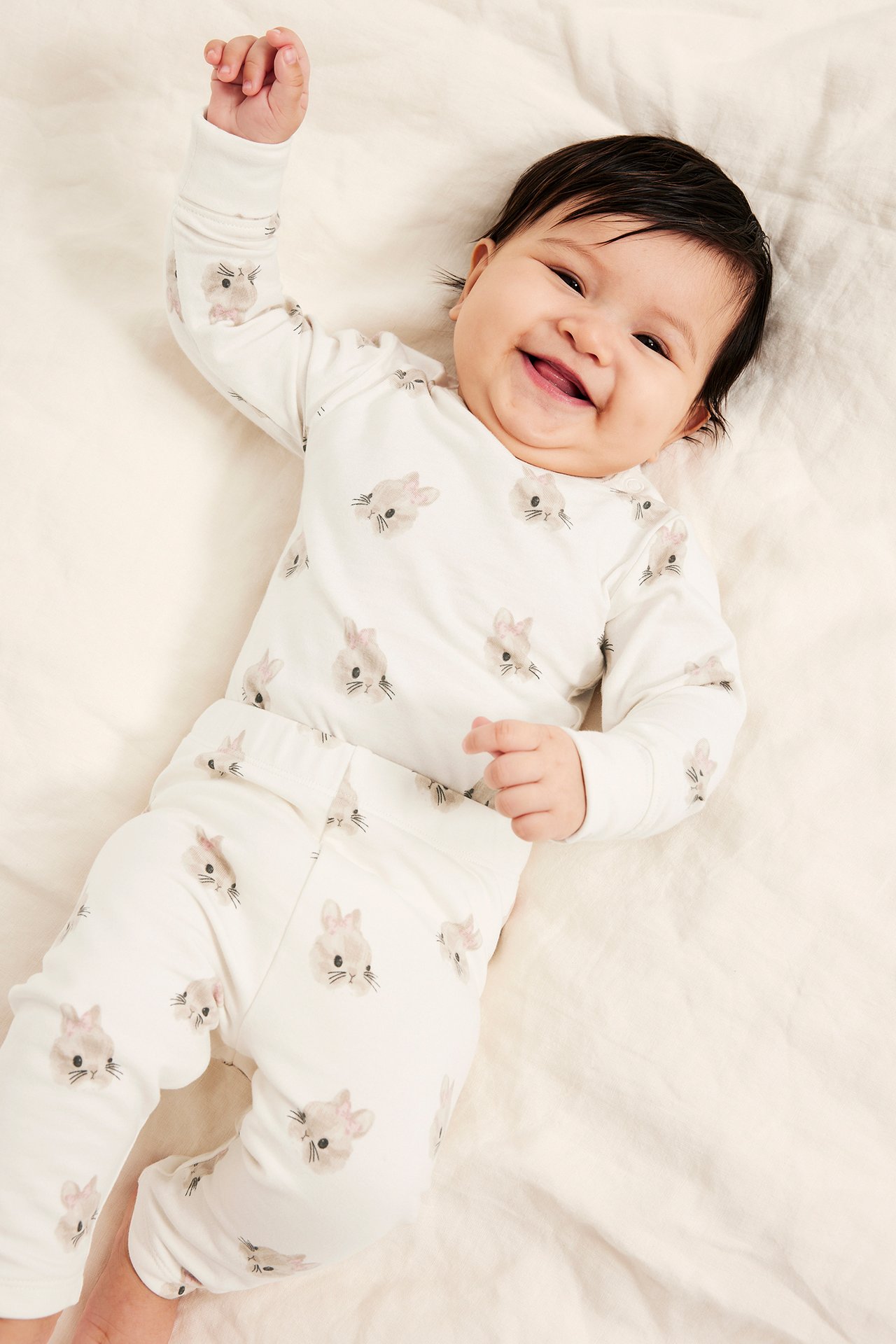 Ylhäältä kuvattu hymyilevä vauva yllään luonnonvalkoinen pitkähihainen body ja siihen sopivat housut pupukuviolla.