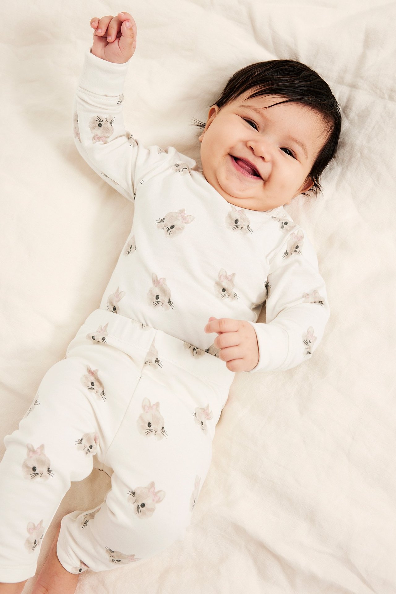 Ylhäältä kuvattu hymyilevä vauva yllään luonnonvalkoinen pitkähihainen body ja siihen sopivat housut pupukuviolla.