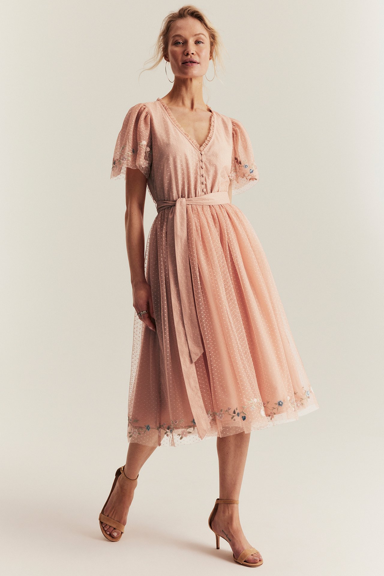 Frontbilde av en kvinne i en rosa prikkete meshkjole med blomsterbroderi, V-hals og beige sandaler.