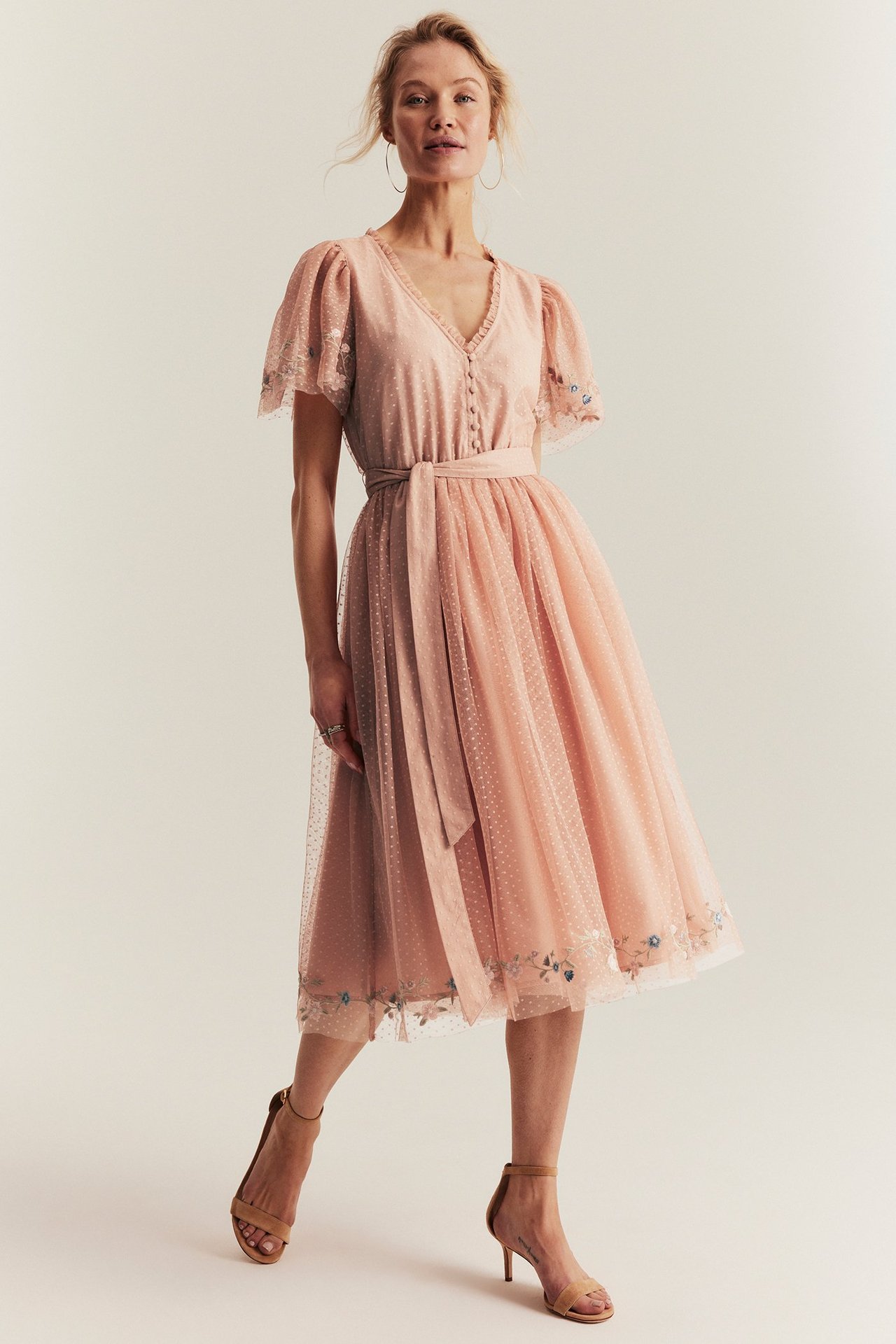 Frontvy på en kvinna i rosa prickig meshklänning med blomsterbroderi, V-ringning och beiga sandaler.