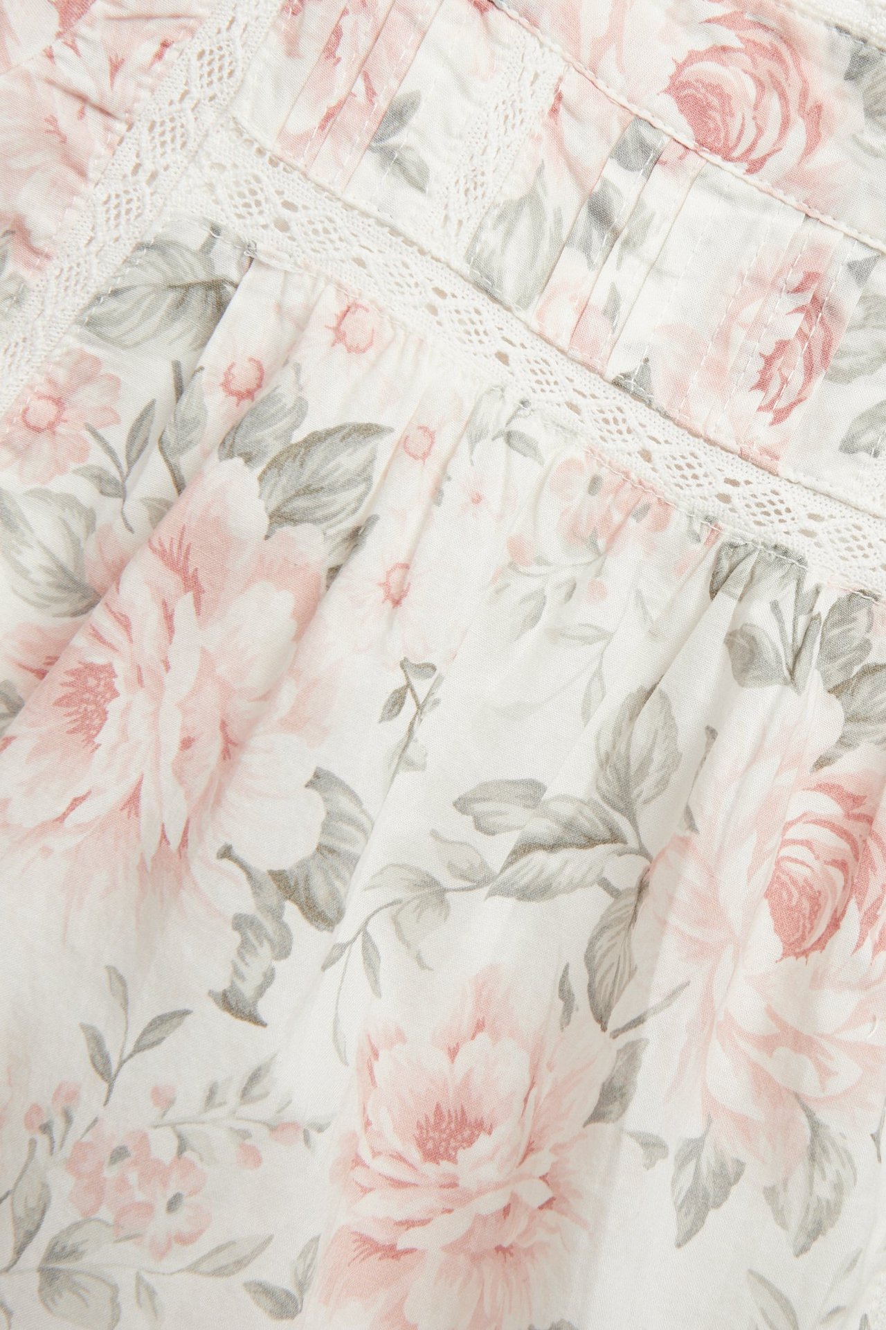 Närbild av offwhite blommig bomullsklänning med rosa rosor, gråa blad, spetsinfällningar och veckdetaljer.
