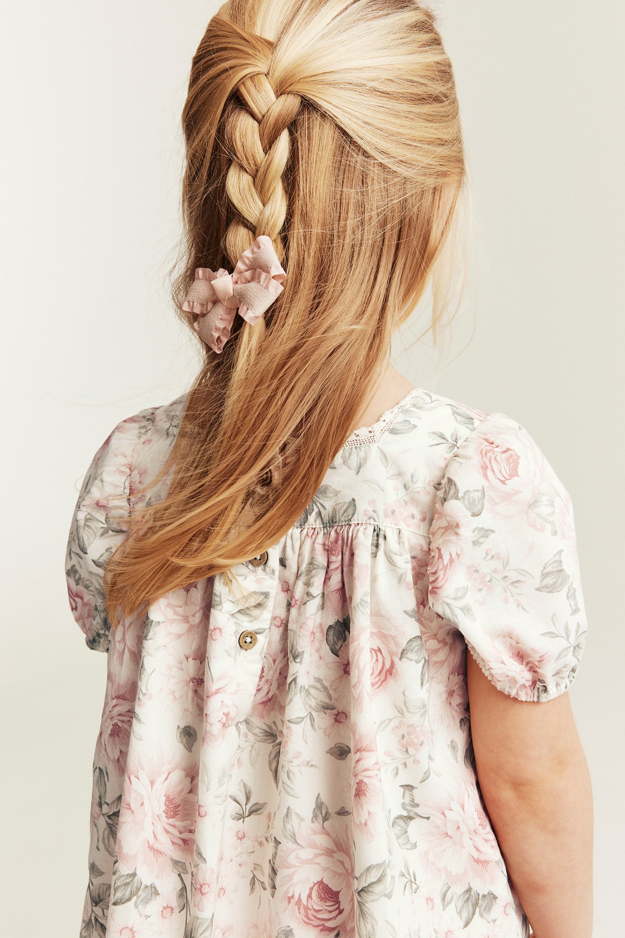 Ryggvy: Barn bär off-white blommig bomullsklänning med ryggknappar, puffärmar. Rosa volangrosett i håret.