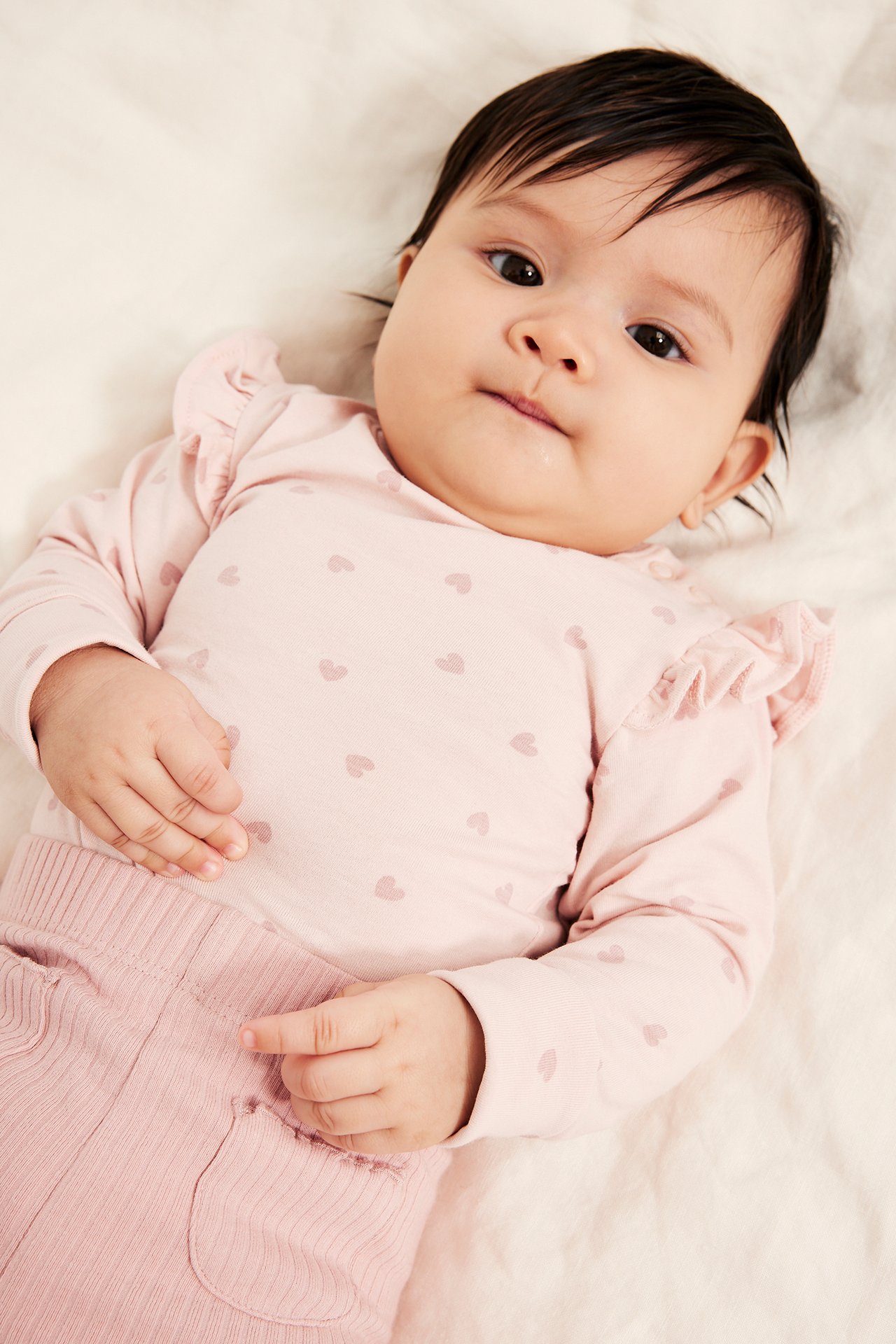 Ylhäältä näkymä: Vauva yllään vaaleanpunainen pitkähihainen sydänkuvioinen body röyhelöolkapäillä ja vaaleanpunaiset ribbihousut.