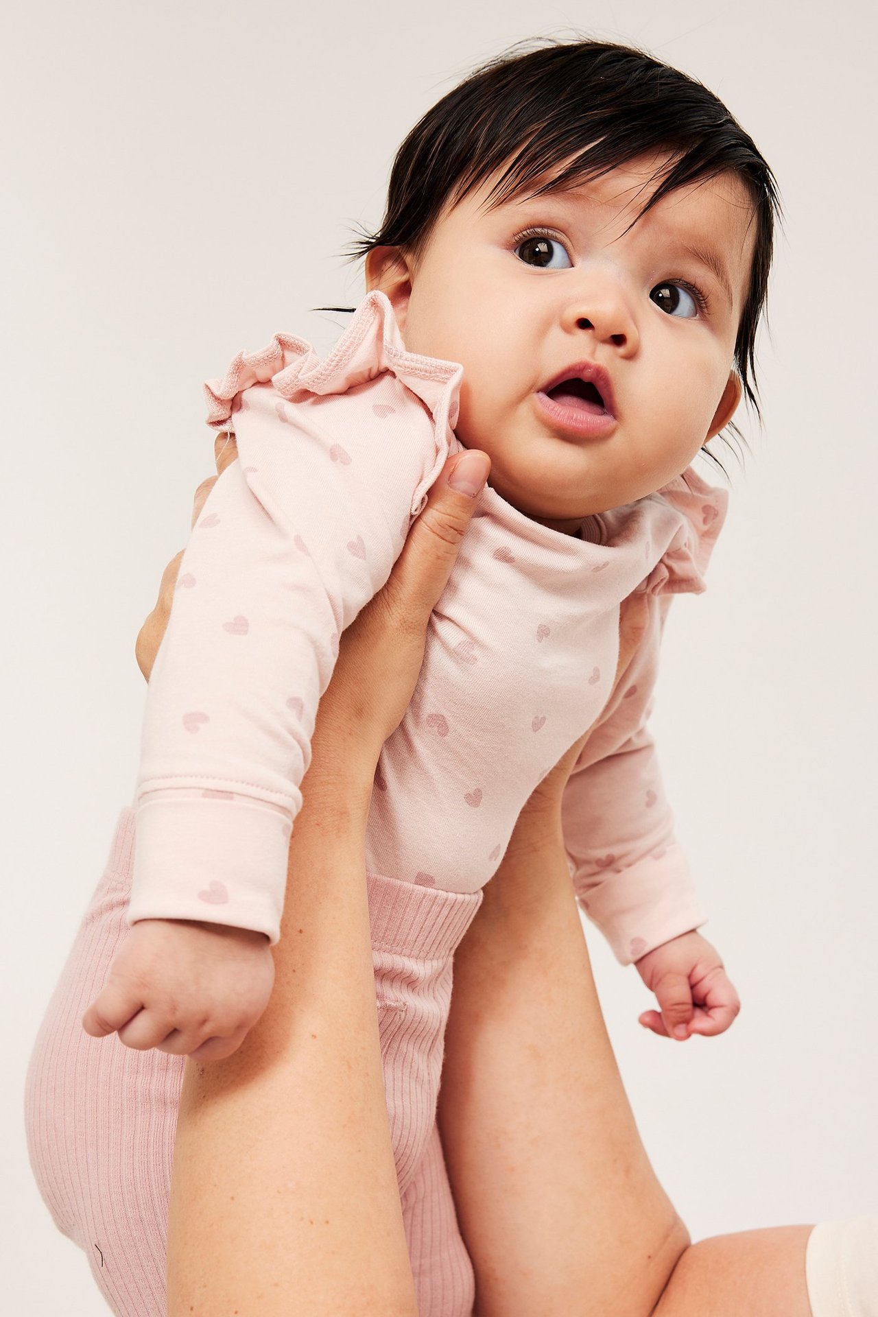 Edestäpäin, vauva yllään vaaleanpunainen pitkähihainen sydänkuvioinen body röyhelöhartioilla ja vaaleanpunaiset ribbineulehousut.