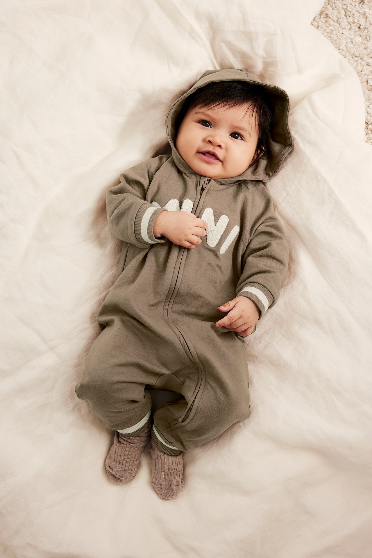 Edestäpäin vauva ruskeassa huppuhaalarissa 'MINI'-printillä, raidallisilla hihansuilla ja ruskeilla ribbisukilla.